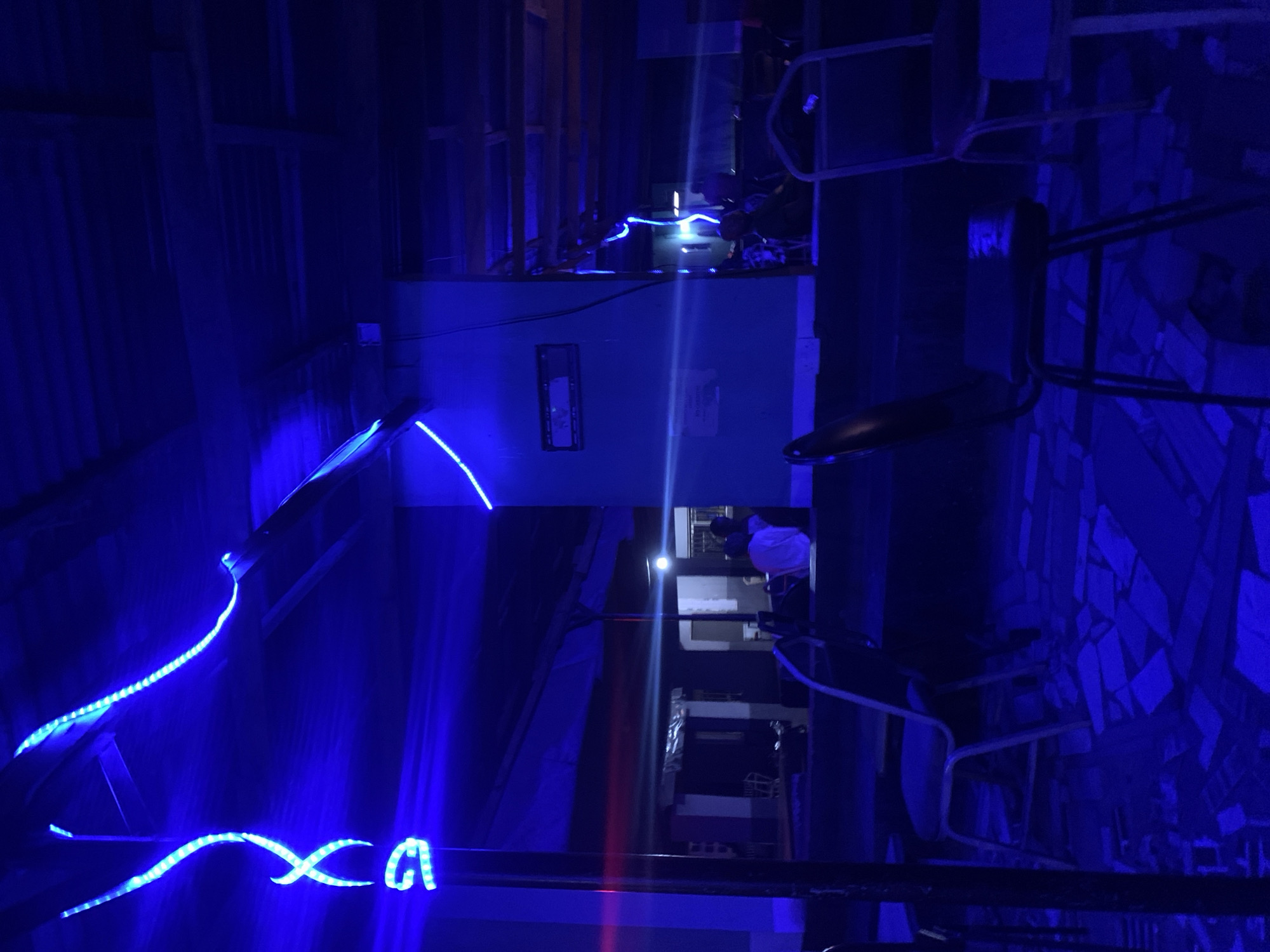 The image depicts a dark, indoor or sheltered outdoor setting illuminated primarily by intense blue LED strip lights, suggesting it was taken at night. The scene is dominated by a cool, deep blue hue.
In the foreground and mid-ground, multiple strands of blue LED lights are visible. One segment of the LED strip on a horizontal bar in the lower left appears to form the stylized letters "xa" or a similar pattern. Another string of blue LEDs snakes along what seems to be a railing or a shelf in the mid-left. A light-colored utility box or cabinet is mounted on a wall in the center.
Structural elements, possibly wooden beams or frames, are visible on the left side of the frame. Towards the right, a wall or surface is covered with irregularly shaped, dark blue tiles. Furniture with metal frames, resembling chairs or stools, is present on the right side of the image. A person wearing a light-colored top is partially visible in the mid-right background. A prominent vertical light streak, likely a lens flare, cuts through the center of the image.
No specific activity is clearly discernible; the scene appears static. The bright blue lighting creates a distinct atmosphere. No other legible text besides the "xa" formation is visible.