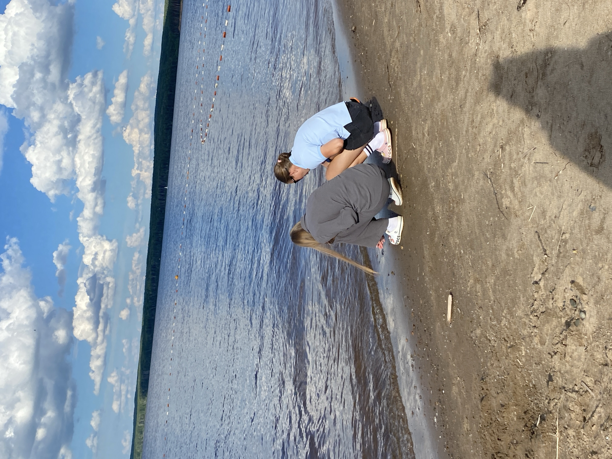 This media file depicts two young females on the sandy shore of a body of water, likely a lake or a river. They are both crouching and appear to be looking at something near the water's edge. The person closer to the water is wearing a light blue t-shirt and dark shorts, with pink and white Crocs. The person further from the water is wearing a grey long-sleeved shirt and dark pants, with white sneakers. Both individuals have their hair pulled back.
The scene is set under a bright, partly cloudy sky, suggesting daytime. The water is calm, with visible ripples on its surface. A line of buoys or markers stretches across the water in the distance, indicating a designated swimming or boating area. The shoreline is sandy and appears to be somewhat wet.
The overall mood seems relaxed and casual, consistent with a day spent outdoors near water. The image was taken in Chaikovsky, Russia. There is no visible text in the media file.