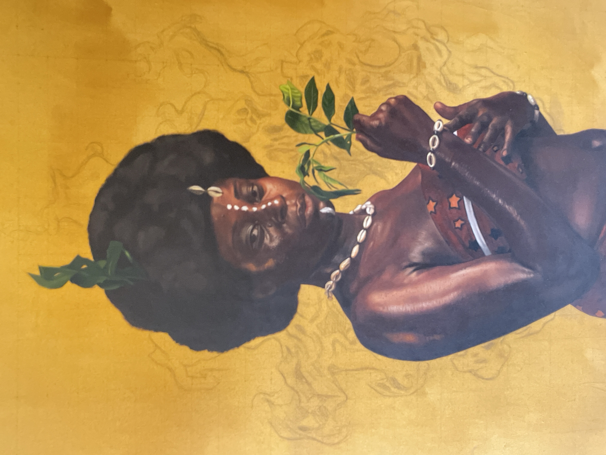 An oil painting on canvas features a Black woman with dark skin and dark brown, afro-textured hair, adorned with cowrie shells arranged in a line on her forehead and as a necklace. She also wears cowrie shell bracelets. She is holding a sprig of green leaves in her hand. Her top features an alternating pattern of orange stars and white stripes. The background is a gold-colored canvas with abstract linear patterns sketched in a lighter shade of gold. The painting may be on display in Ado, Nigeria.