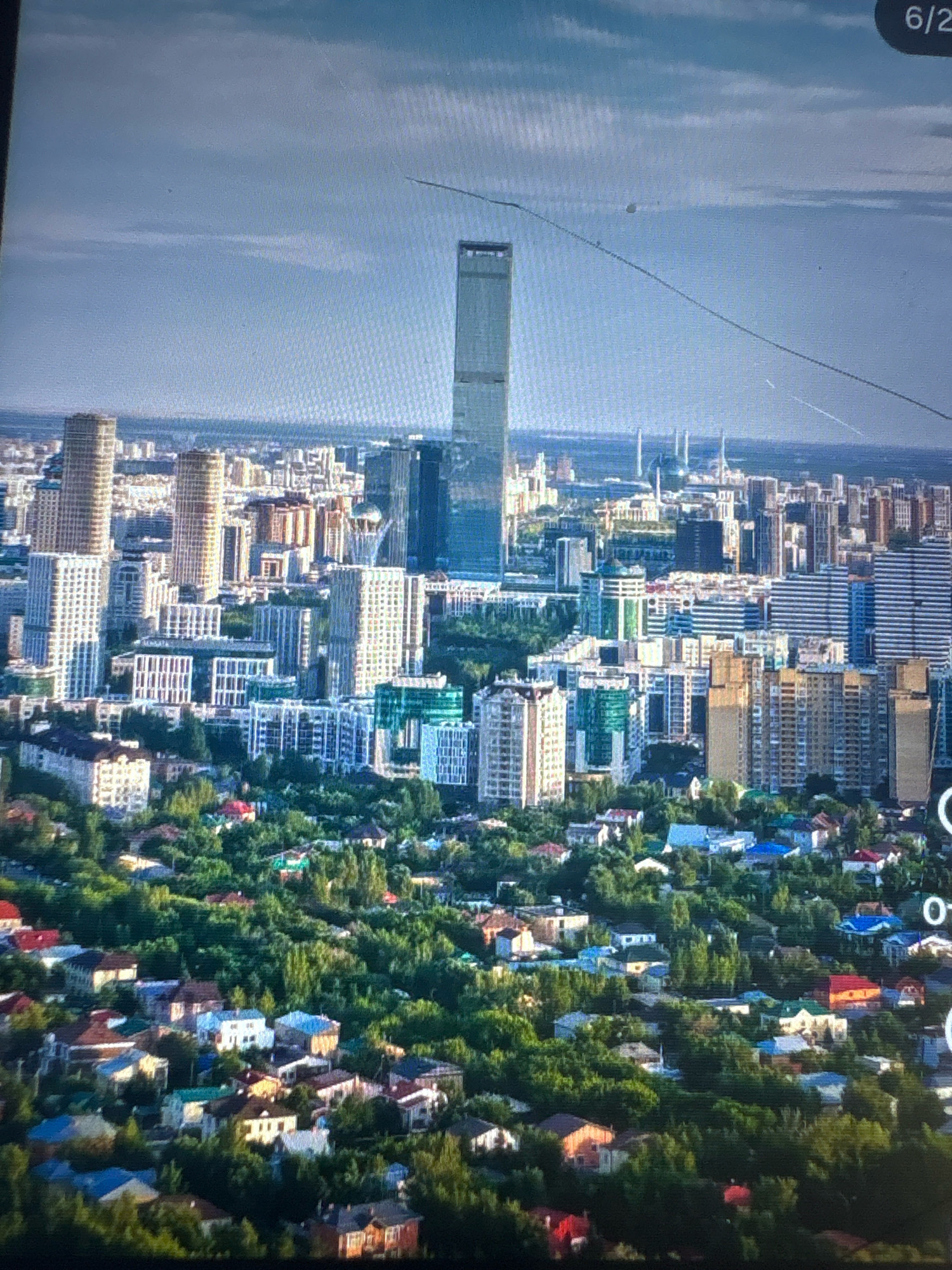 This is an aerial, eye-level, medium shot of the city of Astana, Kazakhstan, with a prominent skyscraper as the central focus. The skyscraper is tall and slender, with a rectangular base and a sleek, modern design. It is surrounded by other buildings of varying heights and architectural styles, creating a dense urban landscape. In the foreground, a residential area with houses and trees can be seen, suggesting a mix of urban and suburban environments. The sky is partly cloudy, and the overall lighting suggests it is daytime. The image is slightly blurry and appears to be taken from a distance, possibly from a drone or helicopter. There are no people or specific actions visible in the image. The overall impression is of a dynamic and growing city with a mix of modern and traditional elements.