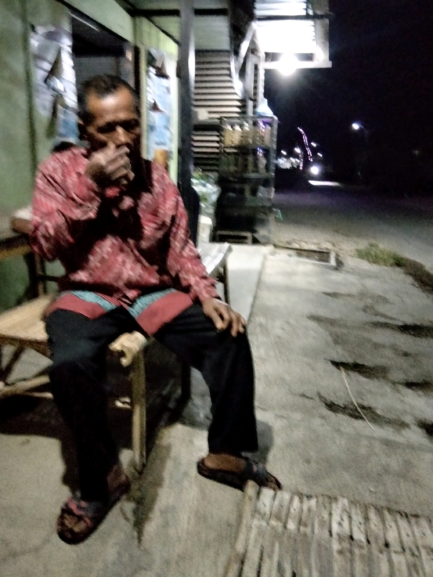 An adult male is seated on a wooden bench outdoors at night in Mrican, Indonesia. He is wearing a red patterned long-sleeved shirt, black pants, and sandals. His right hand is raised to his mouth. The bench is situated on a concrete surface. In the background, there is a building with a light-colored wall and a structure with horizontal slats. The street and lampposts are visible in the distance. The scene is dimly lit, with some streetlights casting illumination.