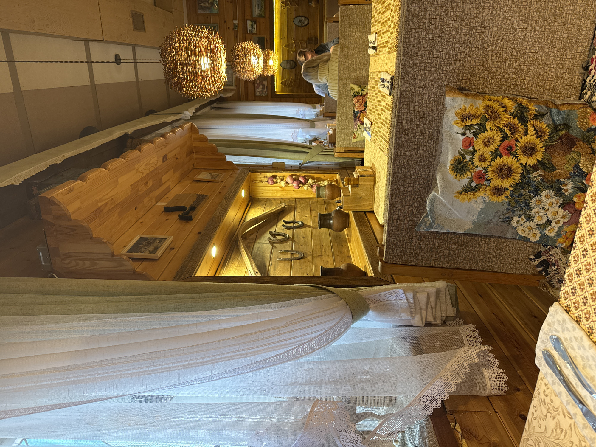 This image captures the interior of a restaurant in Ново-Никольское, Russia, showcasing a rustic and traditional aesthetic. The walls and ceiling are constructed from light-colored wood. Light fixtures made of woven material hang from the ceiling. A wooden display features a collection of earthenware pots, horseshoes, and strings of garlic. Curtains with a delicate lace trim adorn the windows. A table with a patterned tablecloth is set with silverware. A decorative pillow featuring a sunflower design rests on the bench seat. A person is seated at a distant table.