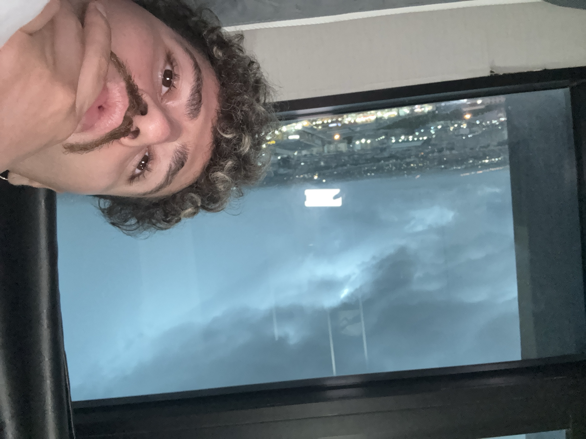 The media file is a selfie taken by a person with curly hair and a mustache. The person is looking towards the camera with a neutral expression. The background shows a window with a view of a city at night, with lights visible in the distance. The sky appears cloudy and dark, suggesting it might be evening or night. The scene is indoors, likely in a room with a window overlooking the city. The person's face is partially obscured by their hand. There is no visible text in the image. The rotation of the image suggests it was taken by holding the camera sideways.