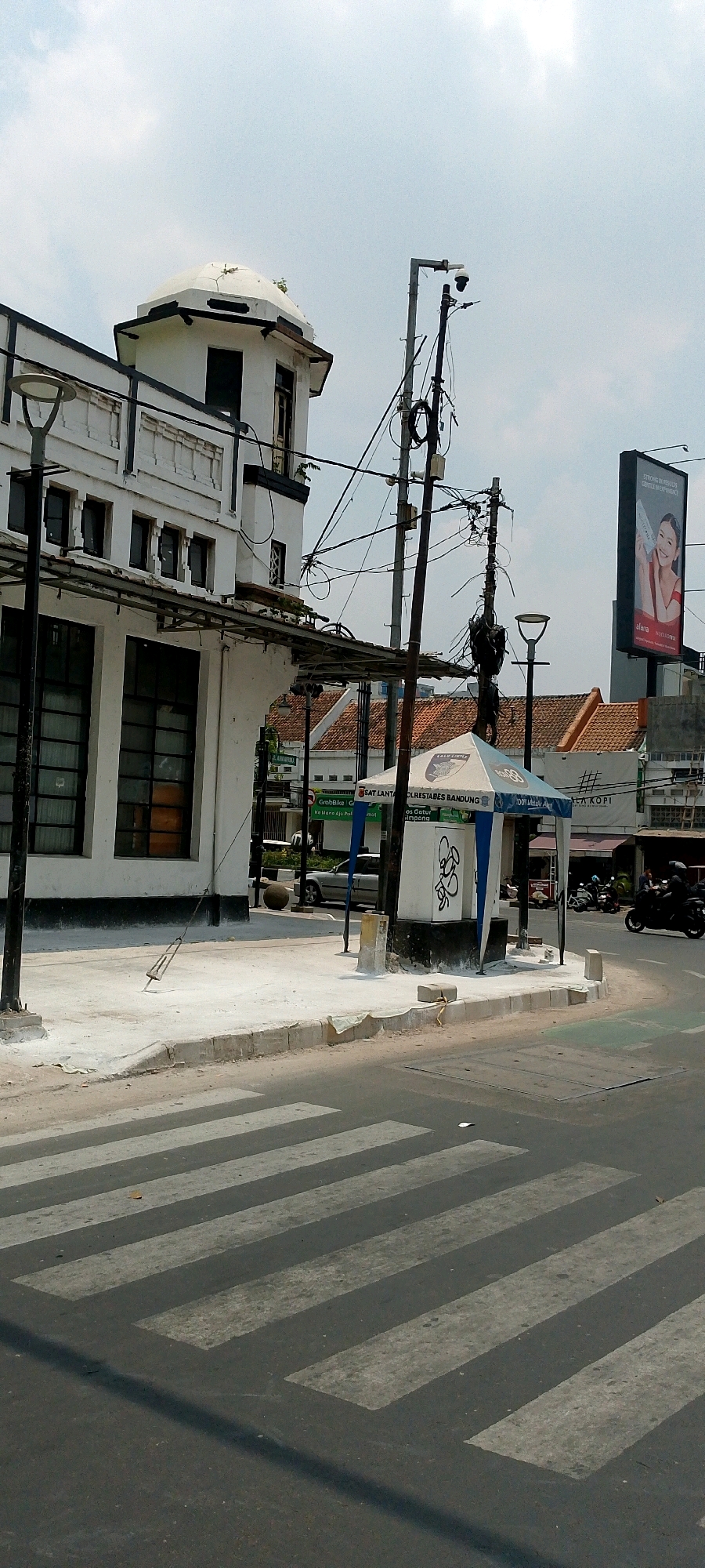 This image shows a street scene in Bandung City, Indonesia, likely near a busy intersection given the presence of a pedestrian crossing and a traffic light pole with multiple cables and a security camera. On the left, a white building with dark-trimmed windows and a distinctive domed structure on top, possibly an older architectural style, stands beside the street. A lamppost is positioned in front of it.
In the center of the image, a blue and white tent-like structure, possibly a temporary information booth or shelter, is set up on a concrete sidewalk. A white utility box with graffiti is visible inside the tent. Several street lights and utility poles with a complex network of electrical wires dominate the midground. Behind the tent, a silver car is parked or driving slowly.
To the right, a large billboard features a smiling woman in a red top, advertising a product. Further back, there are buildings with orange tiled roofs, suggesting a typical urban environment. The street in the foreground has a clearly marked pedestrian crossing with white stripes on dark asphalt. A dark shadow from the utility pole stretches across the crossing. In the background, motorcycles are visible, indicating active traffic. The sky is overcast with some light clouds.