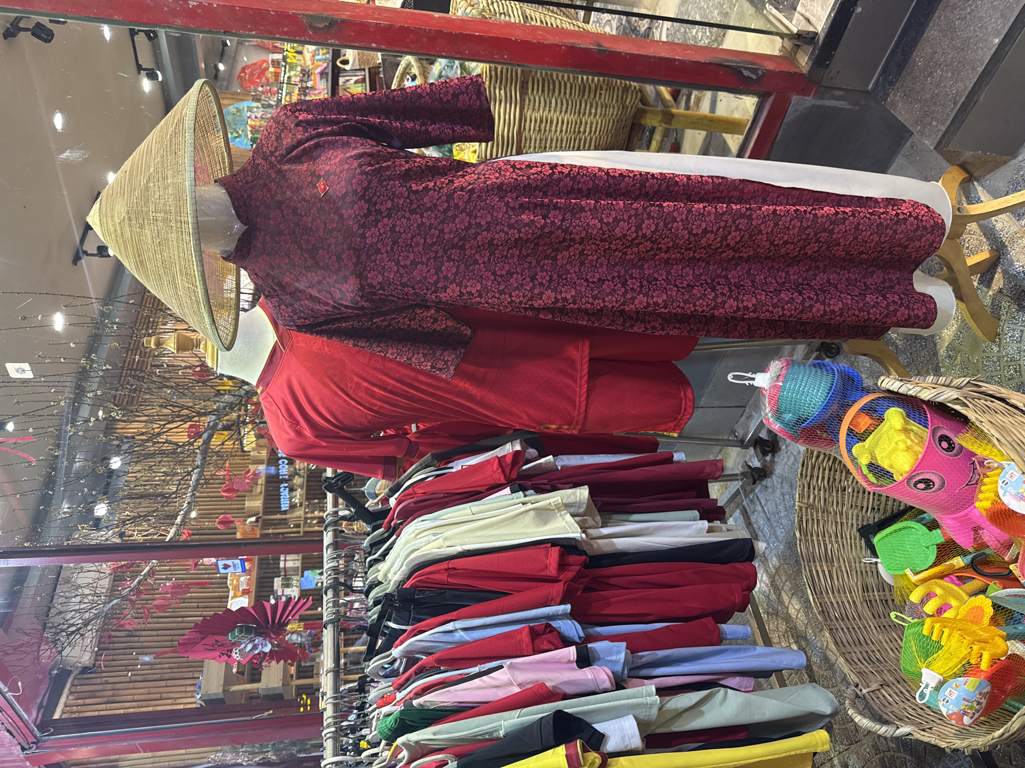 A storefront display in Đà Nẵng, Vietnam features a conical hat atop a mannequin wearing a dark red, floral patterned Ao Dai over a white garment. Below the Ao Dai is a stack of folded clothes in red, white, black, blue, pink, green, and yellow. Next to the clothes, in a wicker basket, are colorful toys. Reflections of bamboo and other red decorations are visible in the storefront window.