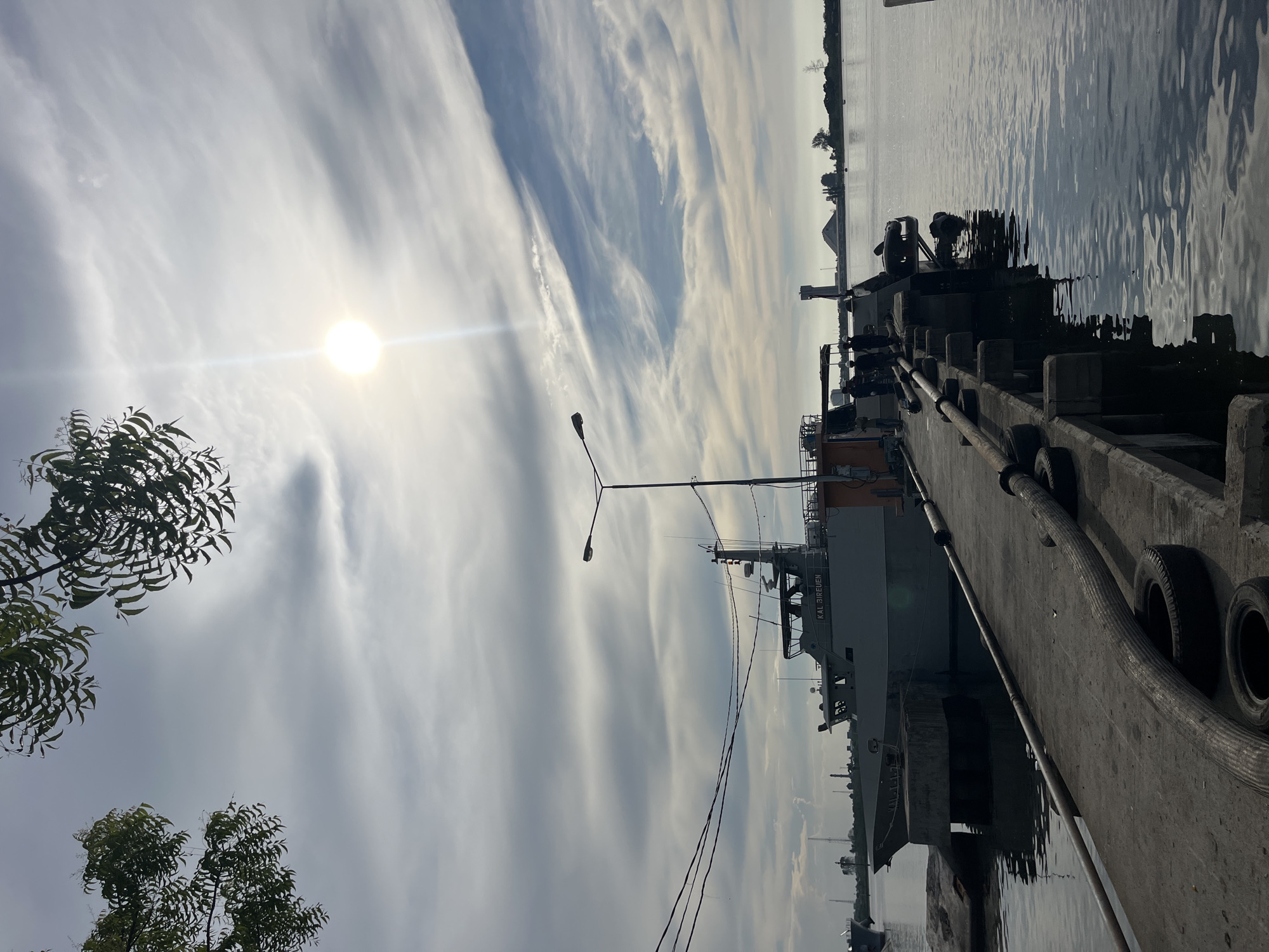 The image depicts a harbor scene in Tambonbaroh, Indonesia. A concrete pier stretches into the water, with a gray ship, labeled "KAL Bireuen", docked along its side. Tires line the pier, likely serving as bumpers. A streetlight stands near the ship. In the background, a body of water reflects the sky, and a few distant buildings or structures are visible. The sky is bright with the sun, and clouds stretch across the frame in long formations. Trees are located in the bottom left. Power lines are also visible.