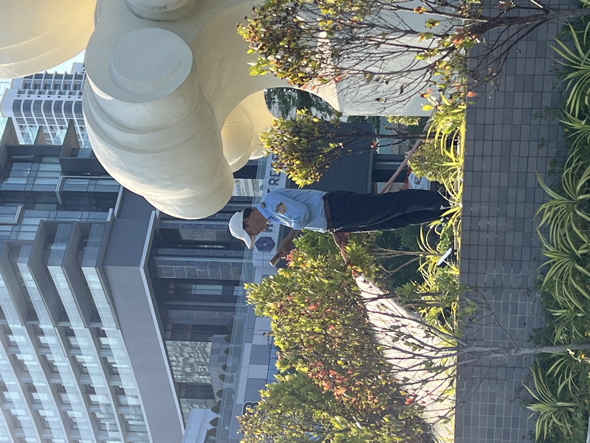A man wearing a white hat and light blue shirt is standing outdoors, appearing to tend to trees or plants. He is positioned in front of a bush or small tree. Behind him is a modern building with a glass facade. To the left, there is a large, abstract, white sculpture. The area is landscaped with trees and plants, and a tiled walkway is visible. Based on the presence of the Cham Towers sculpture, the photo was likely taken in Nha Trang, Vietnam.