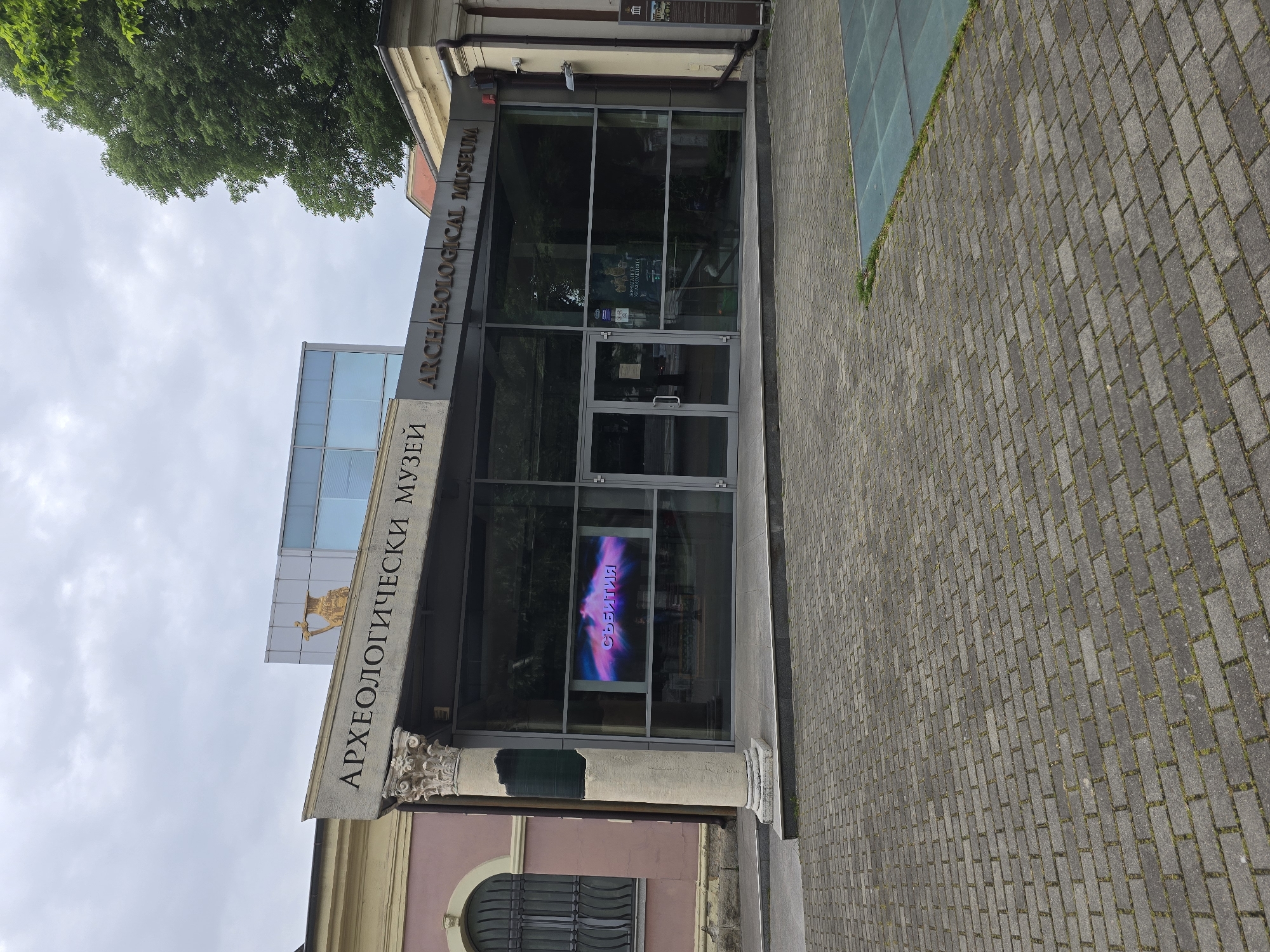 The image shows the Archaeological Museum in the city of Unknown, Unknown. The museum has a modern glass facade with a traditional architectural element featuring a column and ornate carvings on the left side. The name of the museum is displayed in both Bulgarian and English above the entrance. A digital display is visible inside the museum windows. The street in front of the museum is paved with gray bricks. A tree is seen in the upper left corner of the image.