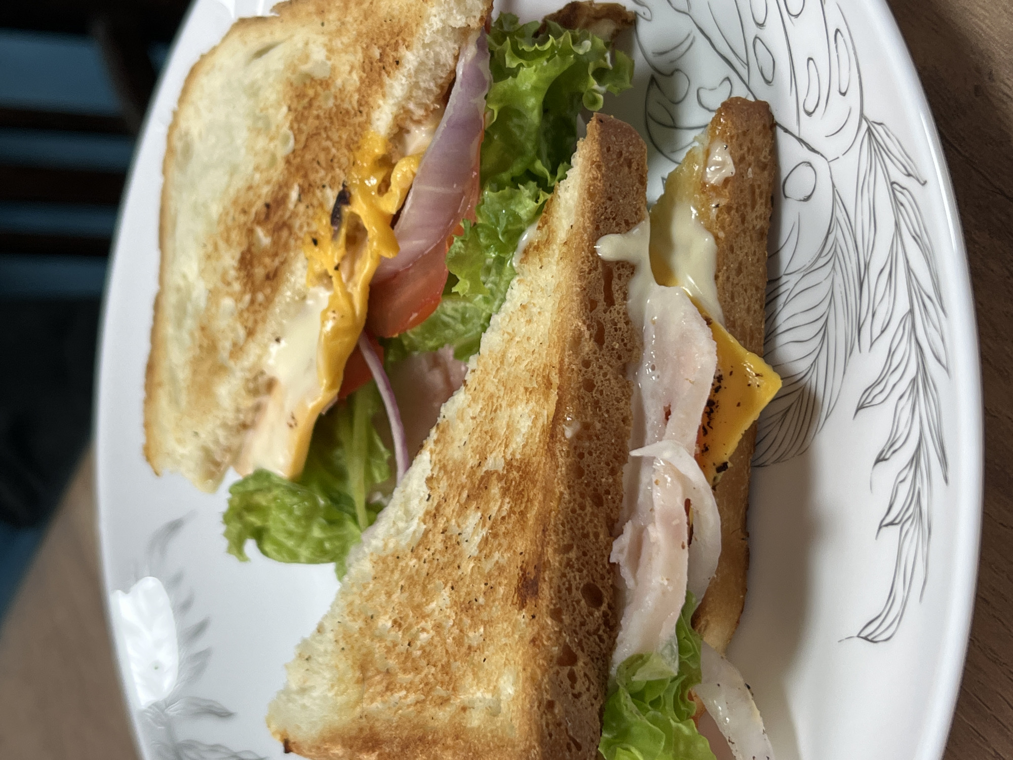 A close-up, eye-level shot shows two halves of a toasted sandwich on a white plate with a faint leaf pattern. The sandwich appears to contain lettuce, tomato, onion, cheese, and sliced meat, possibly turkey or ham, with visible mayonnaise. The lighting is warm and the focus is on the sandwich. The background is blurred, showing a wooden surface and a darker area that suggests furniture. There is no indication of Nizhny Tagil, Russia, or any other specific location.