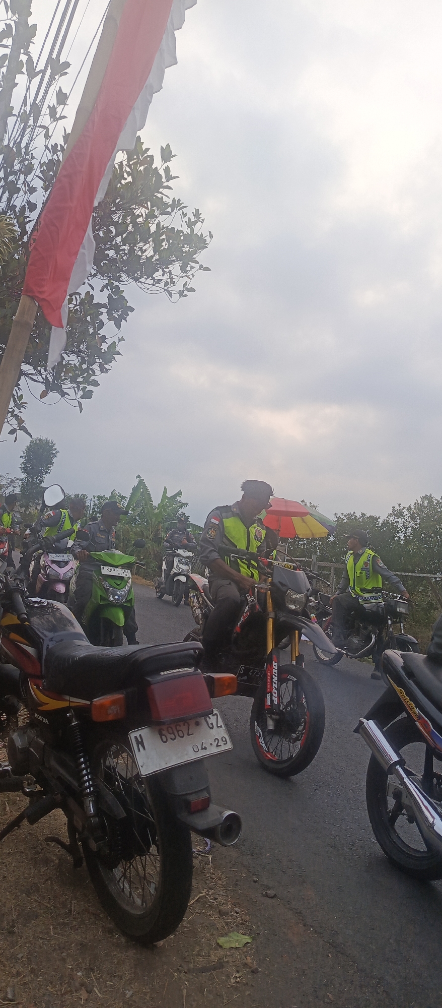 The image shows a group of individuals on motorcycles, some wearing high-visibility vests over their uniforms, suggesting they may be law enforcement or security personnel. The scene appears to be outdoors, possibly on a road in a rural or semi-rural area. A red and white flag, potentially the Indonesian flag, is visible in the upper left corner, attached to a bamboo pole. The sky is overcast, and the lighting suggests it is daytime. Several motorcycles are parked or in motion, with riders seated on them. One motorcycle has a license plate that reads "N 6962 CZ 04-29." There are also trees and foliage visible in the background, indicating a natural environment. The presence of uniforms and motorcycles suggests a possible parade, escort, or organized event. The location, Sumbersekar, Indonesia, is a village in the Malang Regency, East Java.