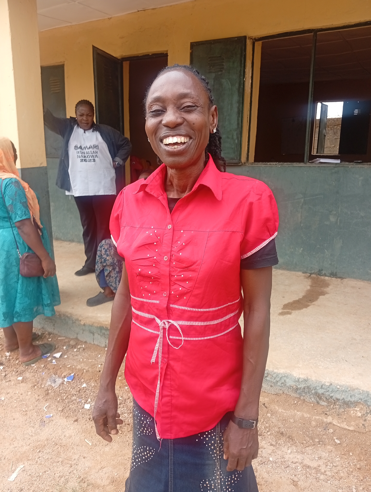 The image captures a vibrant outdoor scene in Narayi, Nigeria, featuring four individuals, with a smiling Black woman prominently in the foreground.
The main subject, positioned centrally, is a woman with braided hair pulled back, wearing a red collared short-sleeved shirt adorned with white decorative stitching and a white tie-belt. She has a dark undershirt with striped trim at the sleeves and a dark denim skirt embellished with white studs. A dark wristwatch is visible on her left wrist. She is looking directly at the camera with a wide, joyful smile, revealing her teeth.
In the background, on a raised concrete step to the left, another Black woman is partially visible. She wears a white t-shirt with the black text "SAMARI IKON ALLAH NAKOWA 2023" printed on it, covered by a dark jacket, and dark pants with dark shoes. She also appears to be smiling slightly. To her left, partially obscured, is a third person wearing a light blue/teal lace-patterned dress, a light-colored head covering, and carrying a small dark brown shoulder bag. Further left, partially hidden by the main subject, a fourth person's legs and feet are visible, wearing dark pants and a patterned garment.
The setting is in front of a simple, single-story building with yellow/ochre-colored walls. Dark green window frames and open windows, some appearing empty, are visible in the background. A dark door frame is also present. The ground is uneven, dusty earth with scattered small stones and debris. The natural lighting suggests it is daytime. The overall mood conveyed by the main subject's expression is one of happiness and positivity.
