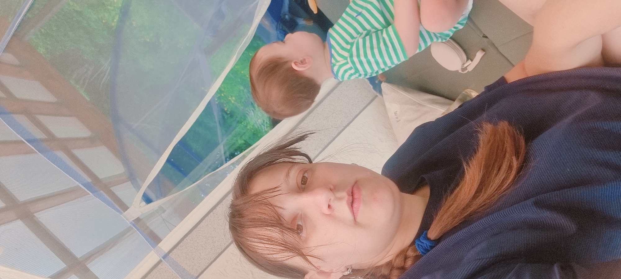 The scene is indoors, likely a room within a dwelling in Dunnae-myeon, South Korea. A woman with light skin, brown hair, and wearing a dark blue garment is visible. She is oriented toward the camera and appears to be reclining. In the background, a child wearing a green and white striped shirt is also reclining. The presence of netting suggests the individuals may be within a mosquito net or similar enclosure. A white object, possibly a headphone or pillow, is near the child. Outside the netting, a green outdoor environment is partially visible.