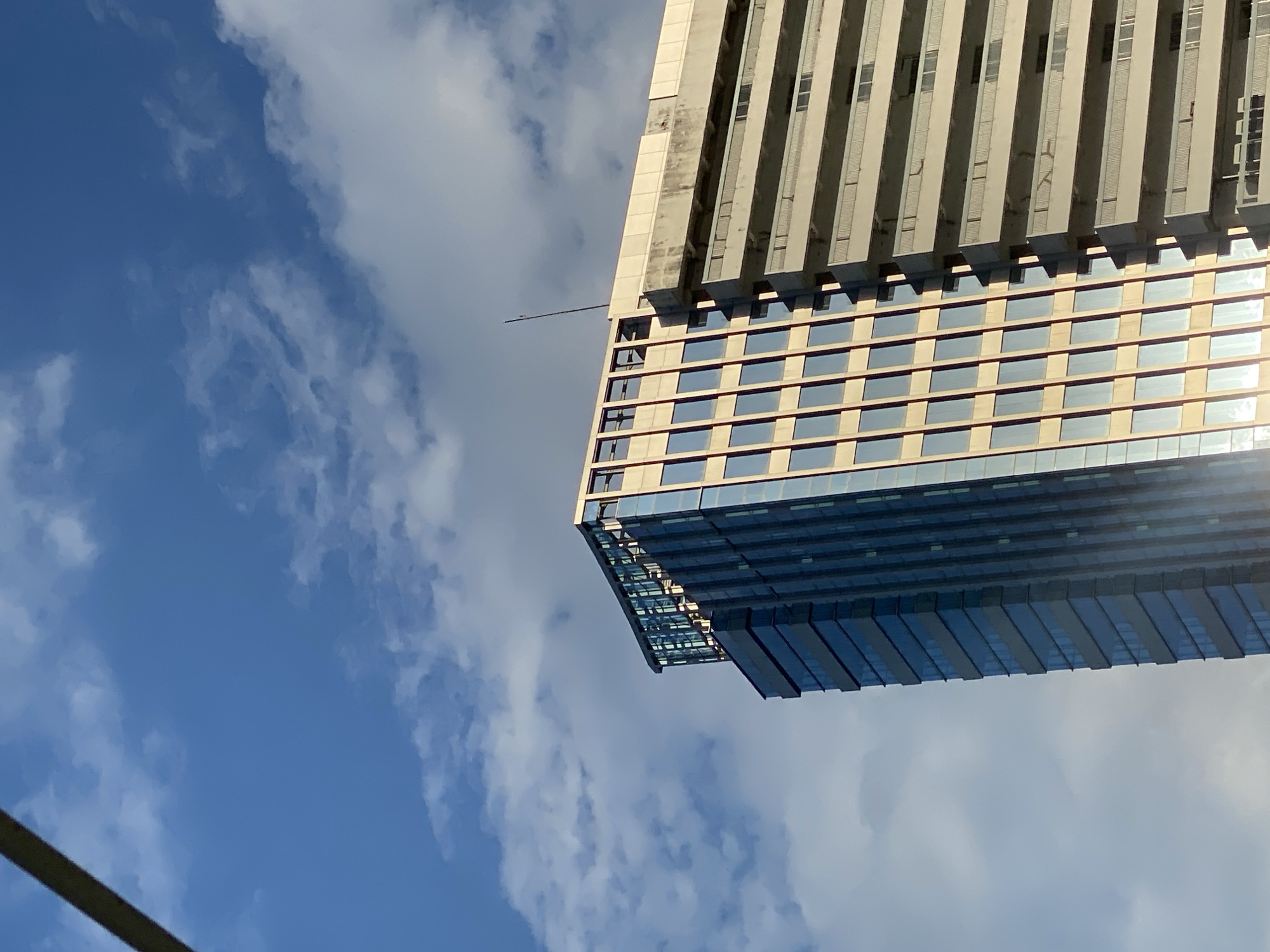 A low-angle shot captures the upper sections of two adjacent buildings against a bright blue sky filled with scattered white clouds. The building on the left is a modern skyscraper with a facade composed of tinted glass windows framed by a grid of dark mullions, creating a sleek and reflective surface. The top of this building features a glass enclosure that catches the sunlight. To its right, a more traditional, lighter-colored building features rows of balconies with railings and numerous windows. A thin, dark antenna rises vertically from the roof of this building. A blurred dark object, possibly a cable or a branch, is visible in the upper left corner of the image, partially obscuring the sky. The lighting suggests it is daytime, with sunlight illuminating the buildings and casting subtle shadows. The scene does not depict any people or specific activities. The location is identified as Gambut Barat, Indonesia.