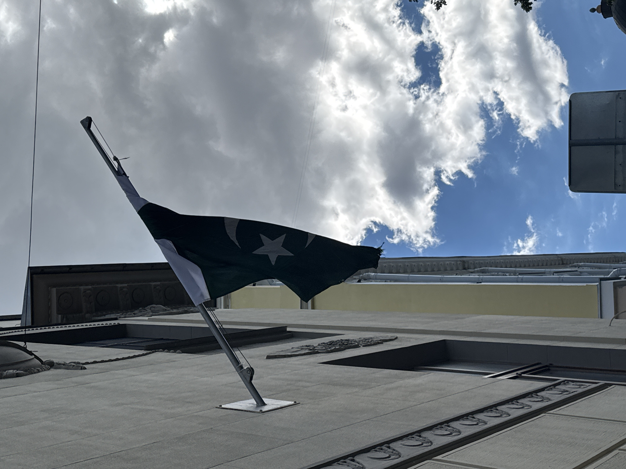 The image captures the Pakistani flag displayed on a building in Moscow, Russia. The flag, dark green with a white crescent and star, is mounted on a metal pole attached to a light-colored building with ornate architectural details. The flag is partially unfurled and appears to be blowing in the wind. Behind the flag, a cloudy sky with patches of blue is visible, suggesting a partly sunny day. The building to the right of the main structure is painted yellow, and a downspout runs along its side. In the lower right corner, a dark dome structure is partially visible. The overall scene is a street view with architectural elements and the national flag of Pakistan prominently featured against a dynamic sky.