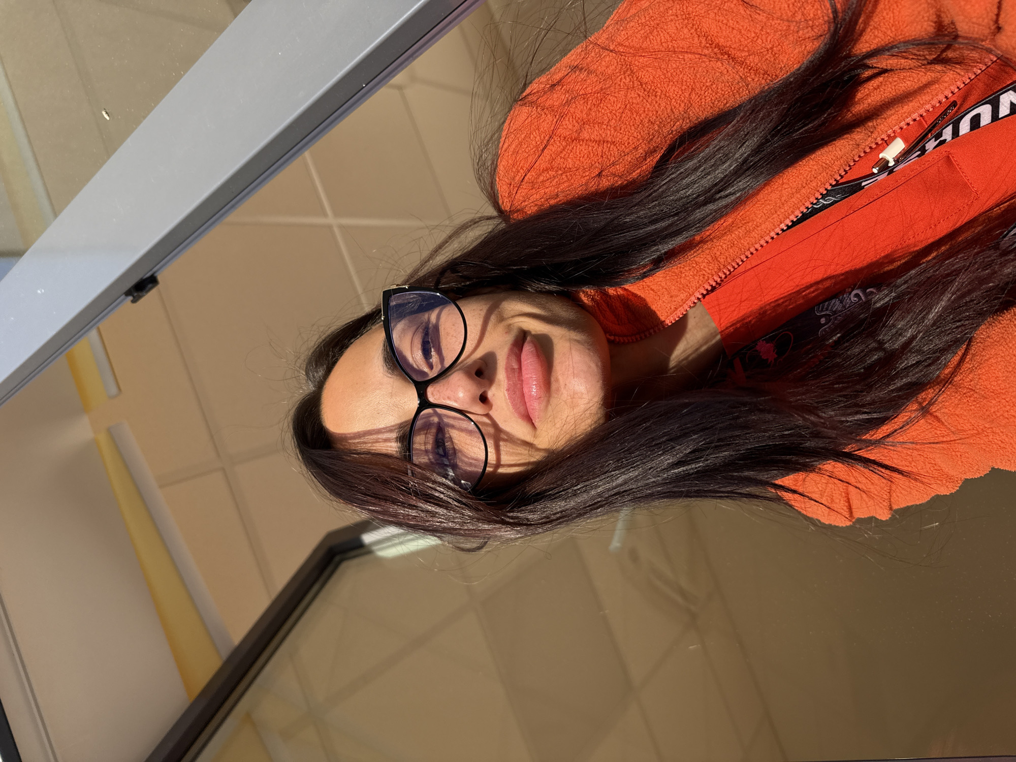 A selfie of a person with dark hair and glasses, wearing an orange zippered jacket, taken at an angle. The background appears to be a reflective surface, possibly a window or mirror, showing a building interior with grey framing. There is no definitive evidence to place this scene in Pleven, Bulgaria.