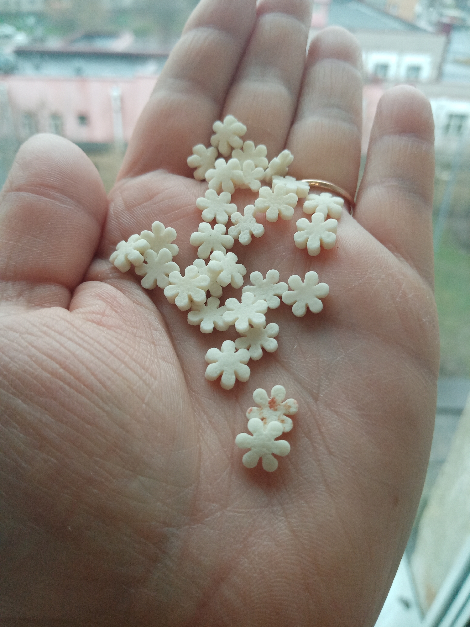 A close-up shows a person's open palm filled with small, white, flower-shaped candies. A gold ring is visible on one finger. Two of the candies have red spots on them. The background is blurry, but suggests an urban scene with buildings and possibly a window. This does not depict the city of Elektrostal, Russia, nor can it be inferred by the provided information.