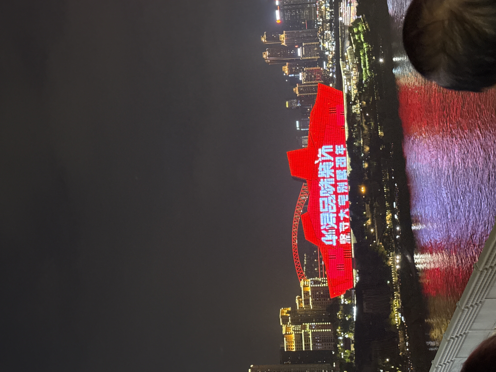 A night time long shot captures a city skyline with a prominent red structure illuminated by LED lights displaying Chinese characters. The structure appears to be a stylized representation of a ship or a building with a ship-like design. The text in red reads "歌唱祖国 坚守初心 砥砺前行" which translates to "Sing the motherland, adhere to the original aspiration, forge ahead." In the background, a cluster of modern high-rise buildings are lit up, and a bridge with a red illuminated arch spans across a body of water. The water reflects the colorful lights of the city and the red structure. The scene is likely located in the Yuzhong District of Chongqing, China, given the distinct cityscape and bridge architecture often seen in that area. A person's head is partially visible in the foreground, suggesting the photo was taken from a vantage point overlooking the scene, possibly from a balcony or bridge.