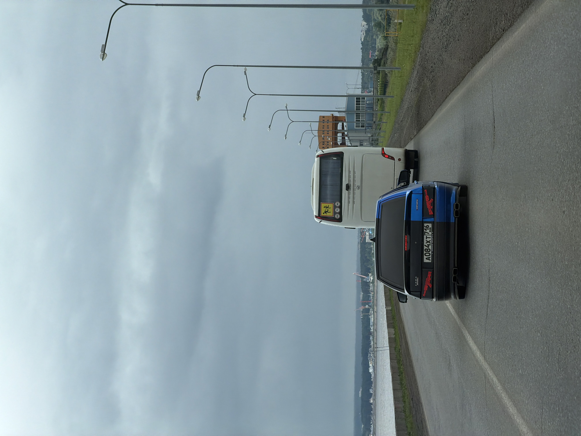 The image captures a road scene in what is identified as Noviy, Russia, under an overcast sky. In the immediate foreground, a modified black and blue Lada Samara car is visible from the rear, featuring a distinct two-tone paint job: black on the left and blue on the right. Its rear lights are customized, with the left light displaying an AK-47 rifle silhouette in red LEDs and the right light showing a red arrow/chevron pattern. The car bears a Russian license plate "A084XT 716 RUS." Immediately ahead of the Lada is a white passenger bus, also viewed from the rear, displaying a yellow square sign on its back window with a black pictogram of two children and speed limits of 60, 70, 80, and 90 km/h.
The asphalt road, marked with a solid white line on its left edge, runs parallel to a large body of water on the left, separated by a concrete barrier. On the right side of the road, a dirt and gravel shoulder leads to a row of tall, modern streetlights with double arms. In the mid-distance on the right, several industrial structures are visible, including one with a distinct orange upper section. Across the expansive body of water in the background, a distant shoreline is apparent, featuring a mix of green vegetation and numerous industrial cranes, suggesting a port or industrial area. The sky is uniformly grey and heavily overcast, indicating a cloudy day. No individuals are explicitly visible in the frame.