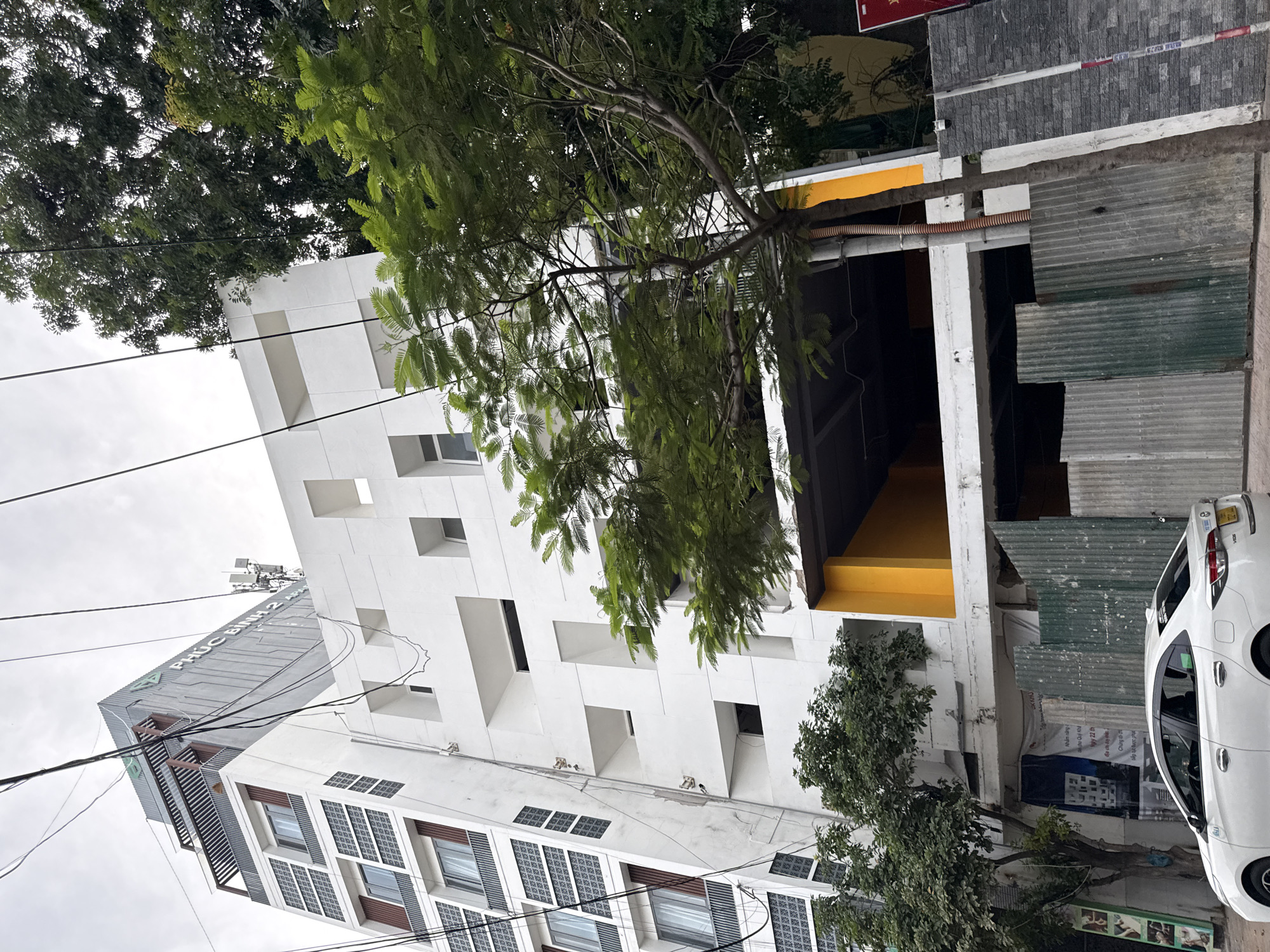 The image shows a street scene in Nha Trang, Vietnam. In the foreground, a white sedan is parked on the side of the road. Behind the car, a section of a building under construction is visible, with corrugated metal sheeting serving as temporary fencing. The building features a bright yellow accent on its facade and has several open levels, suggesting it is a multi-story structure. Above and to the left, a taller building with a grey, textured exterior and the text "PHUC BINH 2" is visible. This building has a modern design with patterned window sections and balconies. Overhead, a dense canopy of green trees partially obscures the sky, which appears overcast. Electrical wires are strung across the frame. The overall impression is one of urban development in progress, with a mix of older and newer architectural elements.