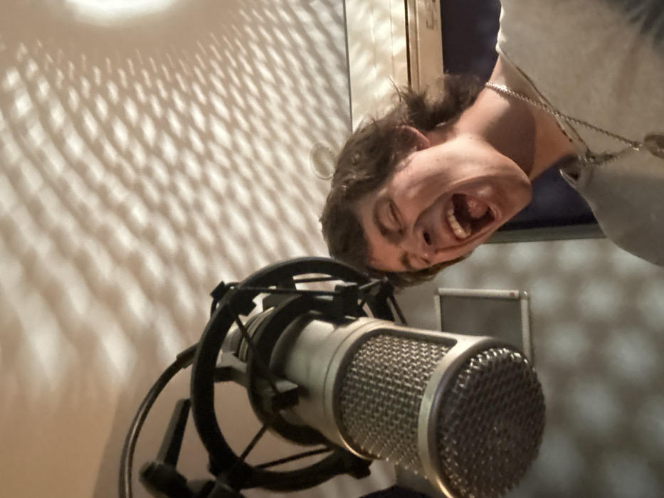 An indoor, angled close-up depicts a male individual positioned in front of a large, professional-grade condenser microphone, mounted in a circular shock mount. The man, with medium-length brown hair, is shown mid-vocalization, his mouth wide open, teeth and tongue visible, and eyes closed, indicating intense vocal exertion. He wears a light-colored, short-sleeved garment and a metallic chain with a circular pendant around his neck. The immediate environment consists of interior walls and a ceiling, with the upper wall/ceiling area displaying distinct, wavy grid-like shadows cast by an unseen light source. A rectangular screen or panel is affixed to a wall section in the background, to the man's left. The scene is consistent with an audio recording session taking place in a studio environment in Paris, France.