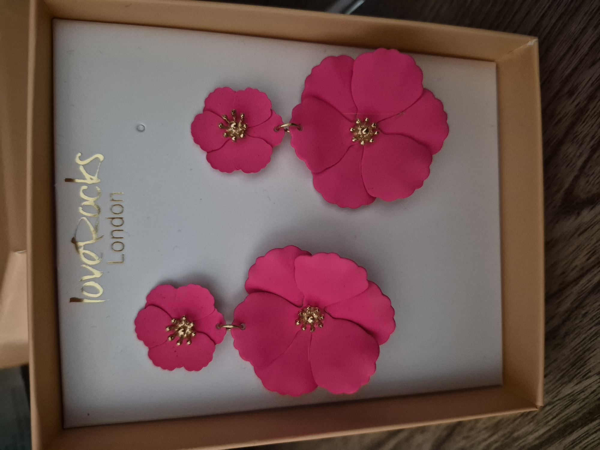 This media file contains a close-up, overhead shot of a pair of bright pink, floral-shaped earrings displayed in a light gray box with a gold border. The earrings are the primary subjects, with two distinct flower shapes on each earring strand. Each flower has five petals and a gold-colored center with small, textured protrusions. The earrings are linked by thin gold chains.
The box the earrings are presented in has the brand name "loverocks" written in gold script on the left side, with "London" written below it in a simpler gold font. The box itself appears to be a light tan or cardboard color. The background is a textured, dark wood surface, likely a table.
The lighting is bright and direct, casting subtle shadows. There is no indication of the time of day or weather conditions. The focus is entirely on the product, suggesting it might be a product shot for e-commerce or a review. There are no people visible, and the setting is contained within the box and the surface it rests on. The overall impression is clean and well-lit, designed to showcase the detail and color of the earrings. The texture of the petals appears to be matte.