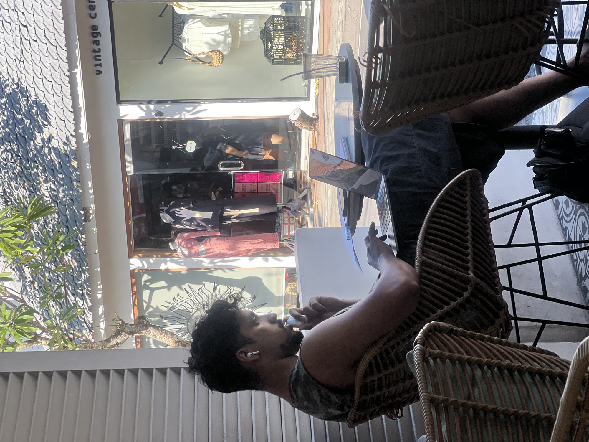 A man with dark hair and a beard sits at an outdoor cafe in North Kuta, Indonesia. He is wearing a camouflage tank top and has wireless earbuds in his ear. He appears to be holding a vaping device. A laptop is open on the table in front of him, with a white piece of paper next to it. He is sitting in a woven wicker chair. A similar chair is visible near his leg, potentially occupied by someone else. The cafe appears to be next to a clothing store called "Vintage Center," based on the sign on the building. The store has mannequins displaying clothing inside. The roof of the building next door features a textured tile design. A small tree is visible near the wall.