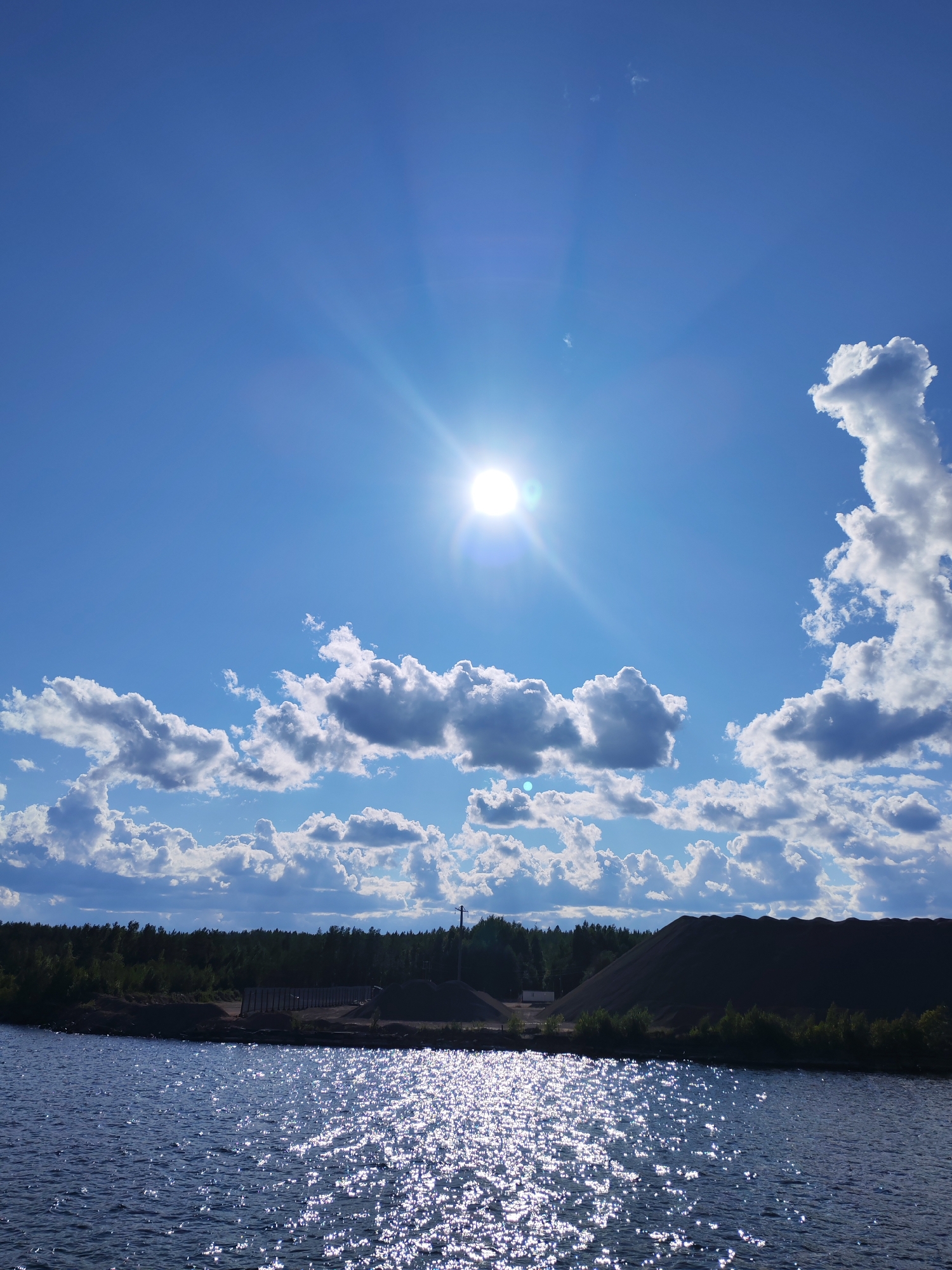 A wide shot captures a bright, sunny day over a body of water and a rural industrial area. The sky is a vibrant blue, dominated by the sun which is positioned in the upper center of the frame, emitting strong rays and lens flare. Scattered white and grey clouds drift across the sky, with one particularly large, wispy cloud formation on the right.
The foreground is filled with the surface of a dark, rippling body of water, reflecting the bright sunlight in a shimmering pattern. Beyond the water, the landscape transitions to a tree line of dark green conifers. In front of the trees, a flat area reveals industrial elements: a low building structure with a fence, piles of dark gravel or dirt, and a tall, slender industrial chimney or tower. A power line pole is visible on the far right. The overall impression is that of a sunlit, industrial waterfront area in Kvartsitny, Russia. There are no people or animals visible in the frame.