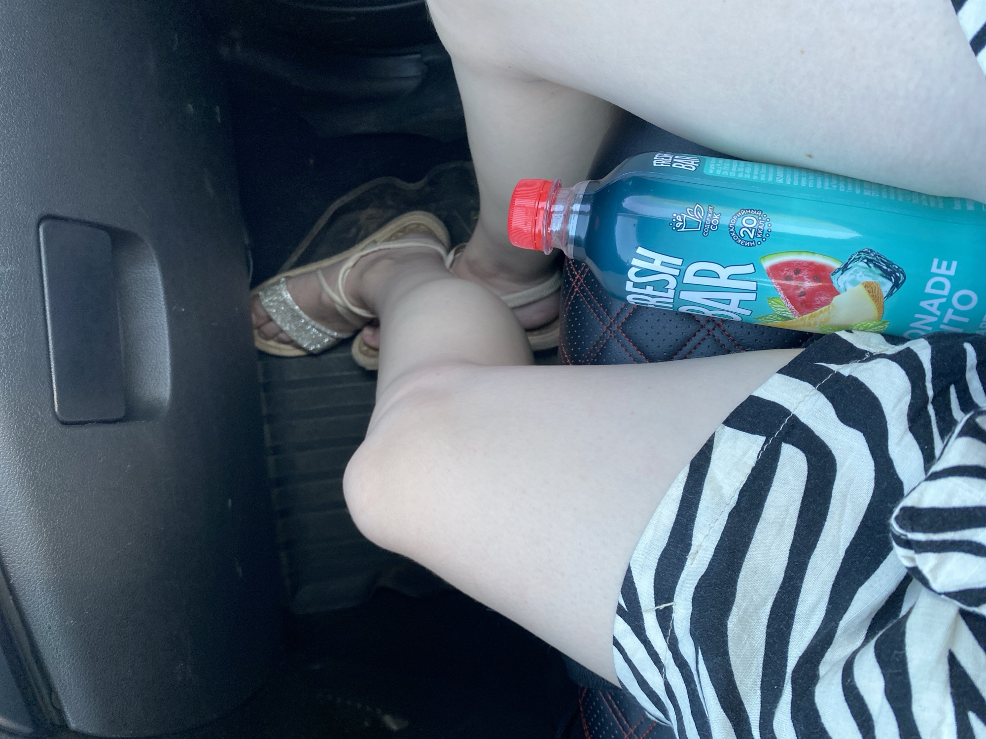A person is sitting in the driver's seat of a car, with their legs crossed. They are wearing sandals with sparkly straps and a black and white striped skirt. A blue bottle of "Fresh Bar" lemonade with a red cap is resting on the person's thigh. The car's interior, including the floor mats and dashboard, is visible in the background. The image appears to have been taken in Kanash, Russia.