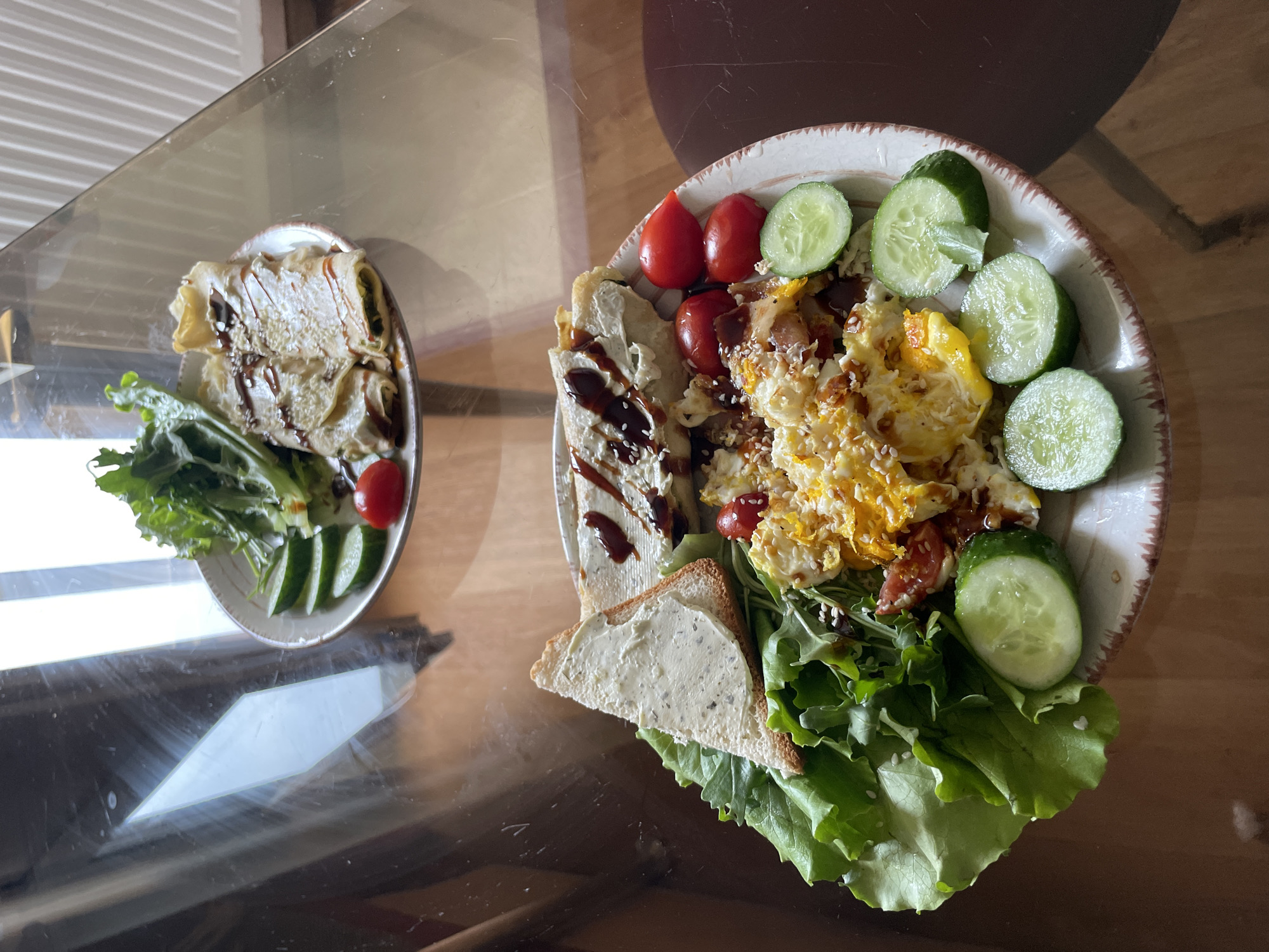 Two plates of food are displayed on a glass-topped table. The plate in the foreground contains scrambled eggs with bacon bits, cherry tomatoes, sliced cucumbers, and a triangle of toast spread with a creamy substance. A rolled-up food item drizzled with a dark sauce and sesame seeds is also present. The plate in the background features a salad with lettuce, sliced cucumbers, cherry tomatoes, and two rolled crepes with a similar dark sauce and sesame seeds. The environment appears to be an indoor setting, likely a dining area, with warm lighting reflecting off the glass table. No people are visible in the image, and there is no specific indication of the city of Kyiv, Ukraine, or any other specific location.