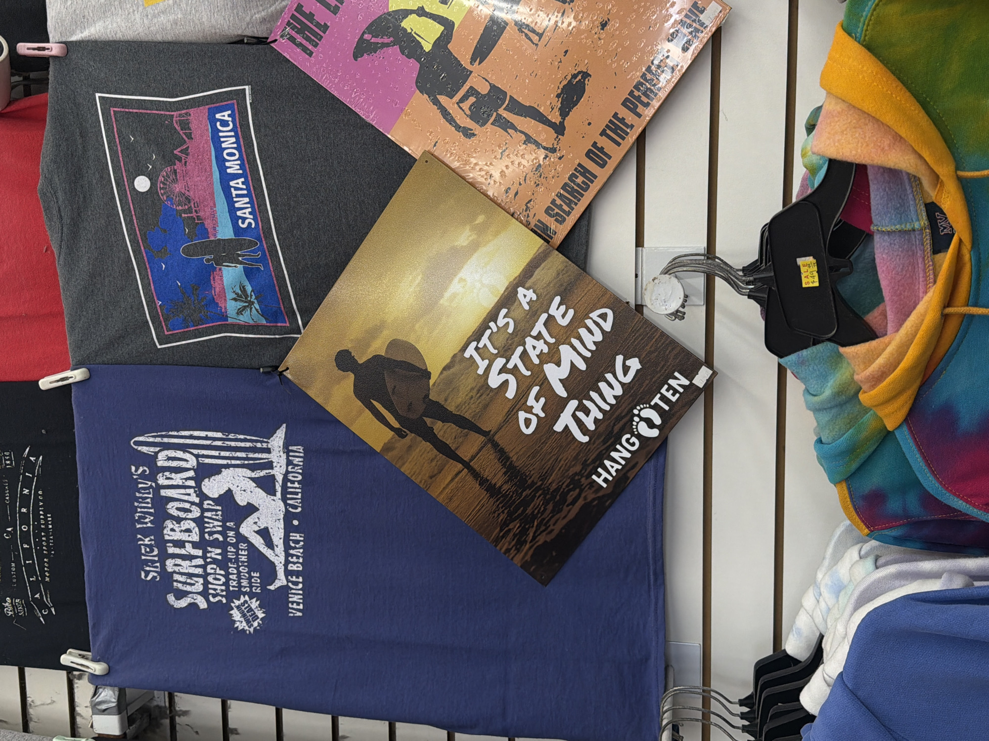 The image displays a retail store setting, likely in Los Angeles, featuring various surf and beach-themed apparel and decorative signs. Multiple t-shirts and metal signs are arranged on a white slatwall display, alongside racks of hoodies.
In the upper left, a dark grey t-shirt shows a graphic of a surfer carrying a board at sunset, with palm trees, a Ferris wheel, and the vertical text "SANTA MONICA". Below it, a navy blue t-shirt features a white graphic of a surfer and a board, with the text "SLICK WILLY'S SURFBOARD SHOP'N SWAP", "TRADE-UP ON A SMOOTHER RIDE", and "VENICE BEACH CALIFORNIA". Partially visible text on this shirt includes "ORIGINAL DESIGNS" and "SINCE 1993". A black t-shirt next to it shows partial text like "Ribo" and "MOTOR SPORT SUPPLY CO.".
Two rectangular metal signs are central. One, with a distressed orange and pink design depicting a surfer, reads "IN SEARCH OF THE PERFECT WAVE". The other, in golden-brown tones, shows a surfer walking into the sunset with the text "IT'S A STATE OF MIND THING" and the "HANG TEN" brand logo (footprints).
To the right, several hoodies are hanging on a rack. A prominent tie-dye hoodie displays vibrant shades of yellow, orange, pink, and teal. A label on one hoodie shows "MV". A price tag on a hanger attached to the tie-dye hoodie indicates "SALE $4.99". The overall scene evokes a casual, surf-centric Californian aesthetic.
