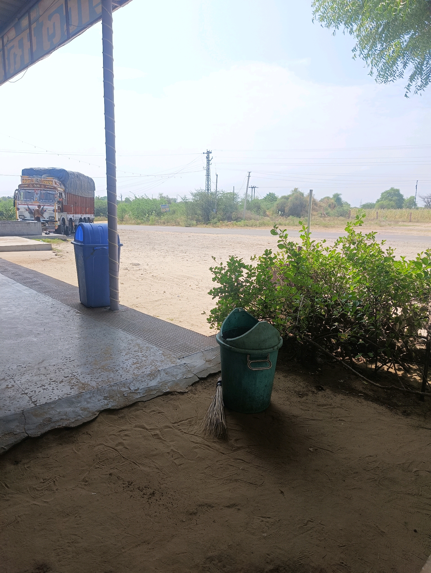 The image depicts an outdoor scene likely near a roadside stop in Chhapri Bari, India. In the foreground, a concrete surface with a metal grate borders an area of sandy ground marked with numerous footprints. To the left, a large blue trash can stands next to a pole wrapped with rope. Further back, a large truck with a covered cargo bed is parked, and a person appears to be near the driver's side. In the middle ground, a paved road stretches into the distance, with utility poles and power lines visible. The background features dry vegetation, scattered bushes, and a bright, clear sky. A green bucket and a broom are visible on the sandy ground to the right of the blue trash can, near a dense cluster of green leafy bushes.