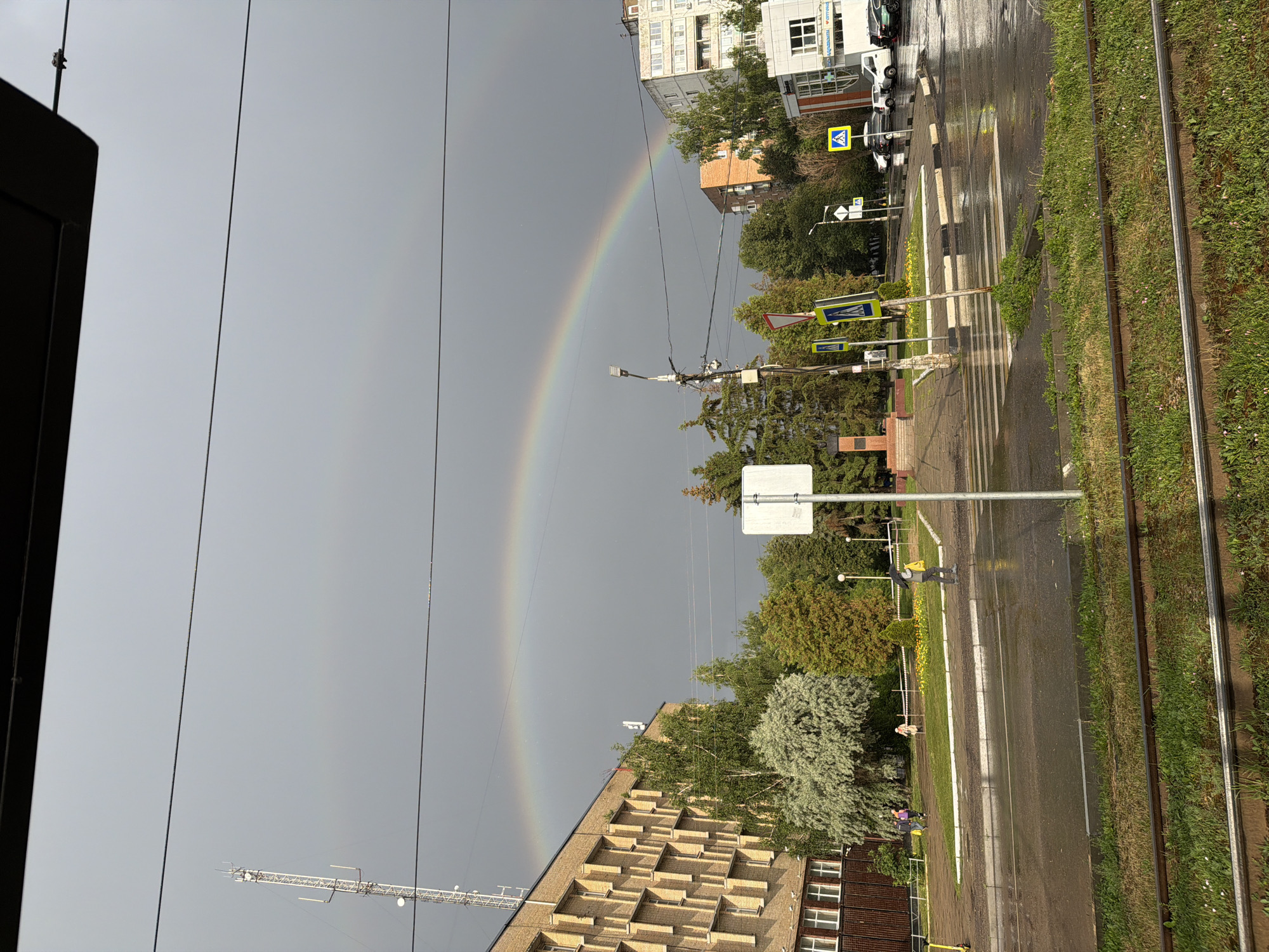 The image depicts a street scene in Kolomna, Russia, under a sky that suggests recent or ongoing rain. A faint rainbow arcs across the overcast sky, indicating a break in the clouds. The street is wet, reflecting the overhead light and the surrounding buildings and greenery.
Several buildings line the street, including a light-colored, multi-story structure with many windows and a darker brick building with a distinctive geometric facade. Trees and foliage are interspersed between the buildings and along the side of the road, adding a natural element to the urban setting.
The foreground shows a portion of tram tracks running alongside a grassy area. A road with a pedestrian crossing is visible, with yellow pedestrian crossing signs placed strategically. A few people are present on the sidewalk and crossing the street, one carrying a yellow bag and another a purple backpack, suggesting movement and daily activity.
The overall impression is one of a damp, post-rain urban environment, with the subtle beauty of a rainbow adding a touch of wonder to the scene. The time of day appears to be daylight, possibly late afternoon or early evening, given the overcast conditions and the angle of the light. No specific text is clearly legible in the image, apart from the signs.