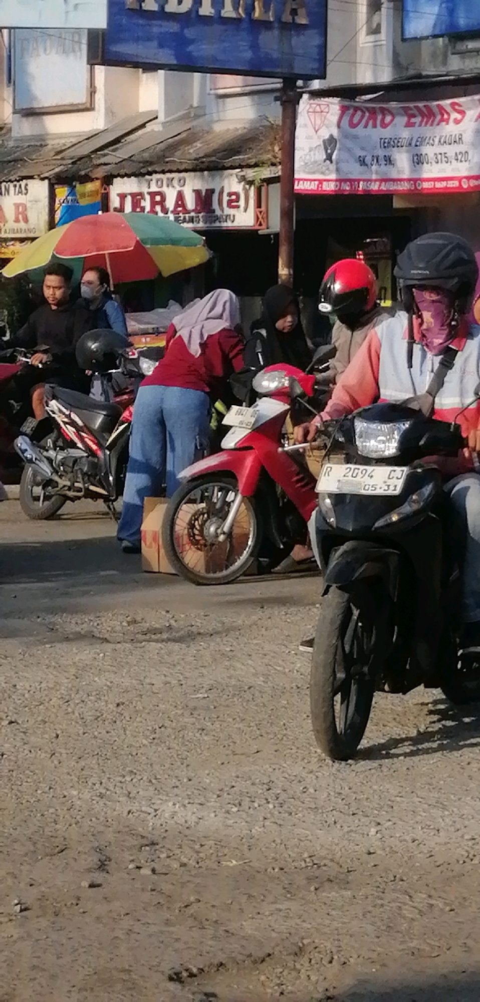The image depicts a street scene in Ajibarang, Indonesia, likely during the day, with several people on motorcycles. In the foreground, a black motorcycle with license plate "R 2094 CJ 05-31" is parked, with its front wheel and lower body visible. Behind it, a red motorcycle is partially seen. To the left, a person wearing a black hoodie is on a motorcycle. Further back, a person in a blue jacket and a face mask stands near an umbrella. Another individual in a red shirt and light blue pants is interacting with someone wearing a light brown jacket and a red helmet, and another person in a black hijab and a purple face covering is also present.
Overhead signs are visible, including one that says "TOKO JERAM(2)" and another that reads "TOKO EMAS". The ground is covered in gravel and some dirt. The overall environment suggests an outdoor marketplace or street setting.