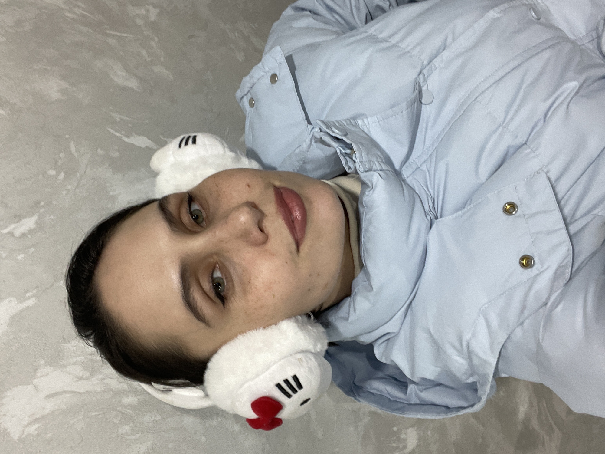 A close-up image depicts a female subject, estimated to be an adult, positioned indoors against a light grey, textured wall with irregular variations in tone. The subject has dark hair, green eyes with winged eyeliner, visible freckles across her nose and cheeks, and light pink lips. She is looking directly at the camera with a subtle smile. She is wearing white, plush earmuffs; the one on her left (viewer's right) is styled with black horizontal lines and a red bow, resembling Hello Kitty. Underneath, she wears a light-colored turtleneck and a light blue puffer jacket with visible silver-colored snap buttons on the collar and front placket. No specific location within Izhevsk, Russia, is identifiable from the visible elements.