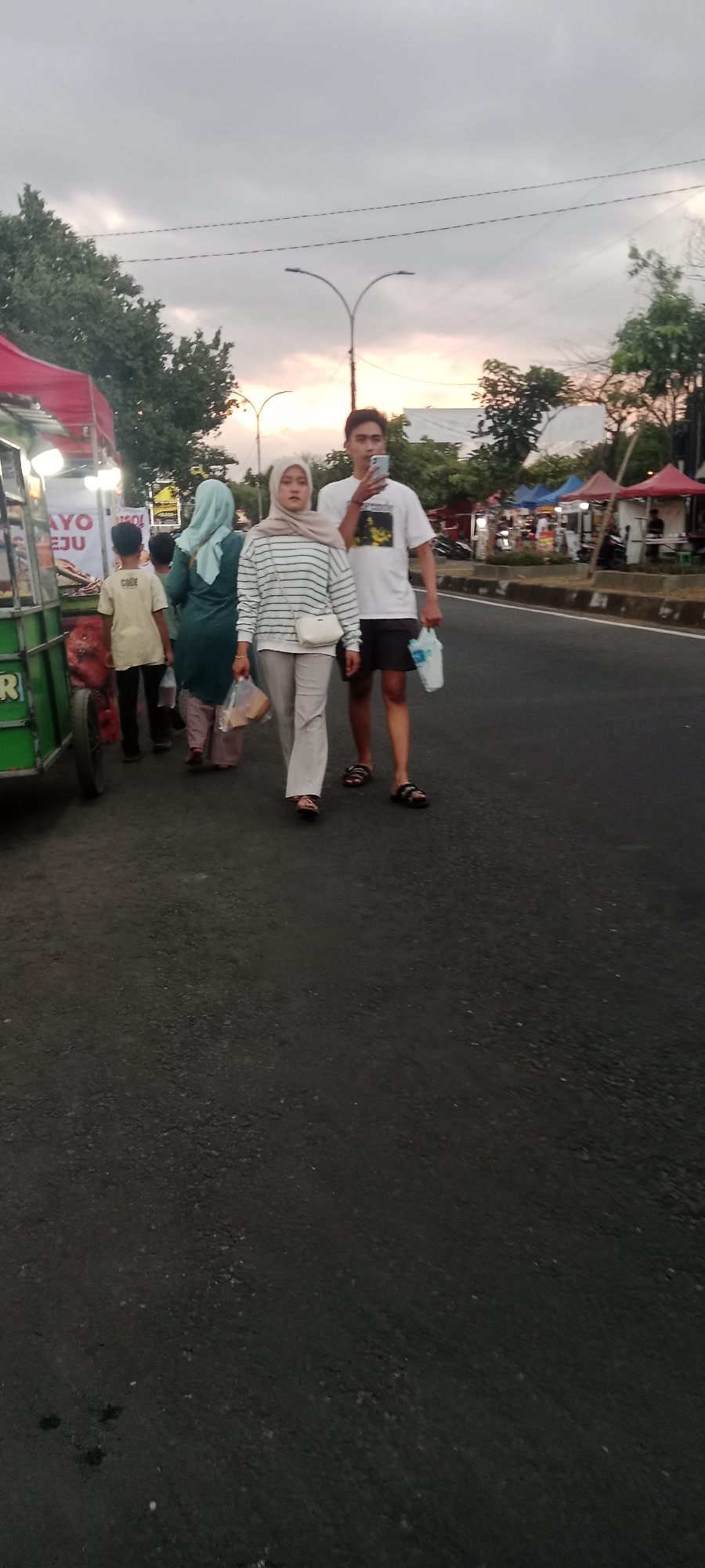 In Ngemplak, Indonesia, a group of people are walking on a paved road during what appears to be dusk. A young man in a white t-shirt and black shorts is holding a smartphone to his face, possibly taking a photo or video, and carrying a plastic bag. Beside him, a woman in a striped long-sleeved shirt and light-colored pants, wearing a hijab, walks with a white crossbody bag and also carries a plastic bag. Behind them, a person in a teal hijab and green dress, and a child in a tan t-shirt and dark pants, walk further ahead. To the left, a food cart with a red awning is visible, with items displayed and illuminated by lights. In the background, there are trees, streetlights, and several market stalls with colorful canopies, suggesting a marketplace or event. The sky is overcast with hints of sunset colors.