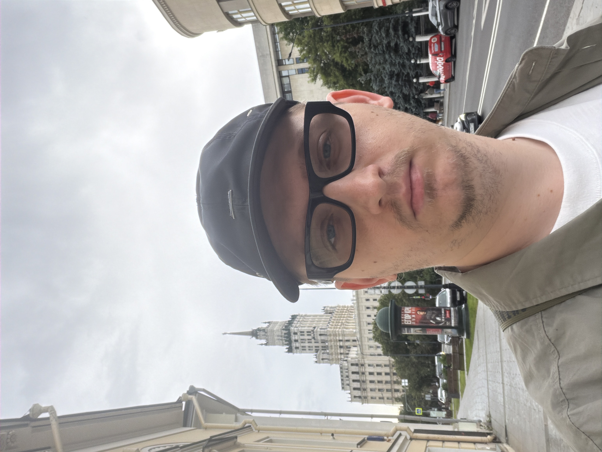 A man wearing a black baseball cap and black-rimmed glasses is taking a selfie outdoors. He has light-colored eyes and a faint mustache and beard. He is wearing a green jacket over a white shirt. In the background, a tall, ornate, white skyscraper with a spire dominates the skyline. The skyscraper appears to be the Kotelnicheskaya Embankment Building, a Stalinist skyscraper in Moscow, Russia. The building is surrounded by trees and other urban structures, including a street with cars and a sidewalk with a poster kiosk. The sky is overcast.