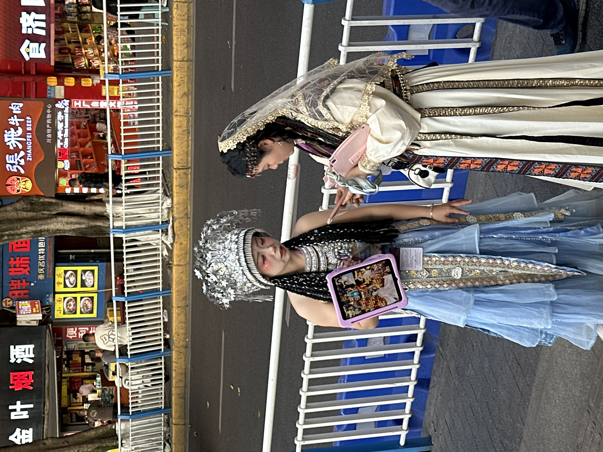 The image shows two women in traditional clothing standing near a blue and white barricade, likely a safety barrier along a street. The woman closest to the camera is wearing a silver headdress, elaborate jewelry, a black braided wig, and a blue dress with layers of fabric. She is holding a tablet with images on its screen. The other woman is wearing a white dress with patterns, a veiled headpiece, and is holding a pink purse or tablet.
In the background, there are storefronts with Chinese signage. One sign reads "ZHANGFEI BEEF" and another mentions "Chongqing" and "Xiao Mian". The shops seem to be selling various products. The storefronts suggest this is a commercial area. The location is likely the Yuzhong District of Chongqing, China, based on the presence of the Chinese signage and the storefront information.