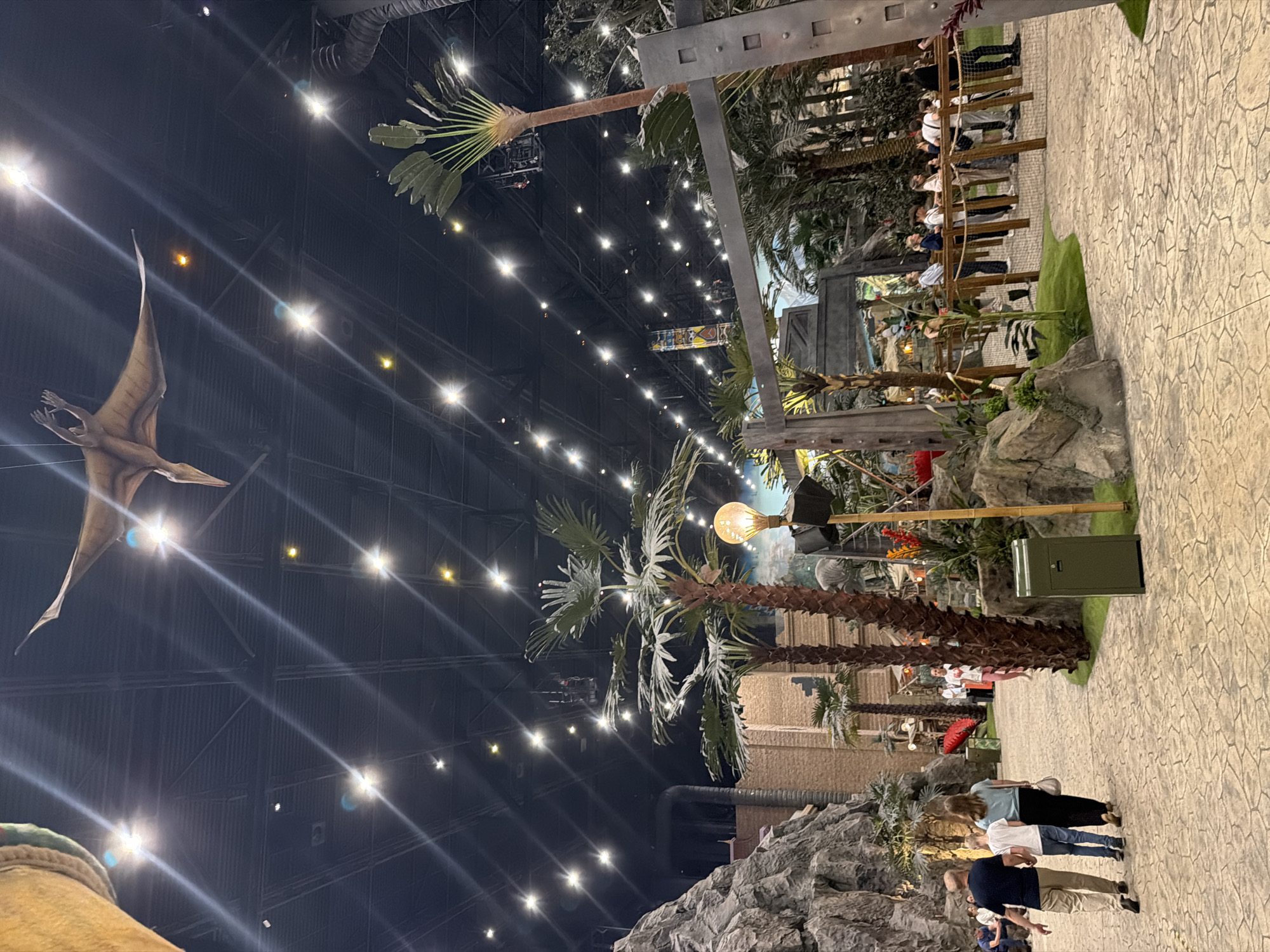 This media file depicts an indoor exhibit with a dinosaur theme. The scene features a large, overhead view of a cavernous space with a high, dark ceiling adorned with numerous lights, creating a starry effect. A prominent subject is a life-sized replica of a Pterodactyl model, suspended from the ceiling as if in flight, its wings spread wide.
The exhibit is designed to resemble a prehistoric landscape. Several tall, artificial palm trees with large fronds are scattered throughout the space, some appearing to be made from rough, textured material. Rocky outcrops and faux vegetation are also visible, adding to the naturalistic illusion. In the background, brick structures and industrial elements of the building's interior can be seen, hinting at the exhibit's construction within a larger venue.
A group of people are present, viewed from a slightly elevated perspective. They are seen walking along a paved pathway, which has a textured, stone-like pattern. The people are casually dressed, suggesting a leisure activity or visit to an attraction. One group of three individuals, including a woman and two men, are walking away from the camera. Another group of people are seen further in the distance, standing near a railing.
A single, large light bulb, encased in a wire cage, hangs from a pole near some of the palm trees, providing localized illumination. A green trash receptacle is also visible on the paved area. The overall impression is of a dimly lit, immersive environment designed for entertainment and educational purposes, likely a museum or theme park attraction. The time of day is irrelevant as the scene is indoors, and the weather is not a factor. No visible text is present in the image.