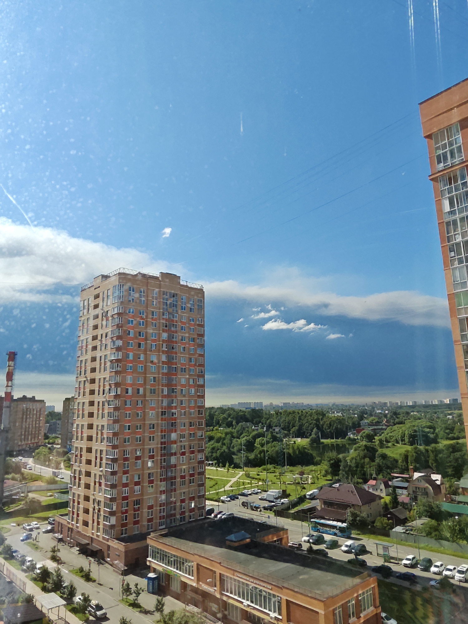 The image captures a cityscape with a bright blue sky dotted with scattered white clouds. In the foreground, a tall, light-brown residential building with numerous windows and red accents stands prominently. To the right, the corner of another orange-brick building with large windows is visible. Below the buildings, a street is lined with parked cars and trees. In the middle ground, a sprawling green area with more trees and houses can be seen, suggesting a suburban or park-like setting within the city. In the distance, a hazy skyline of more buildings extends across the horizon. The scene appears to be a contemporary urban environment. Based on the context provided, this scene is located in the city of Shcherbinka, Russia.
