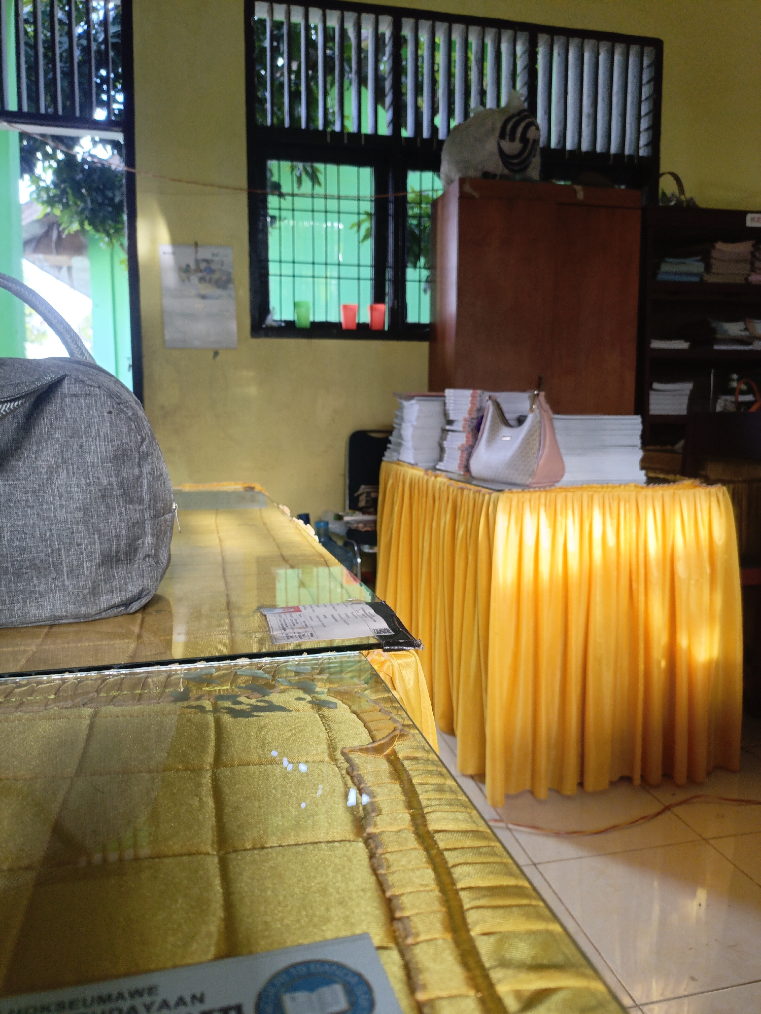 This image captures an indoor scene, possibly a classroom or office, within Lhokseumawe, Indonesia. The room is brightly lit, with sunlight filtering through a barred window on the left, illuminating the space and casting shadows. The walls are painted a pale yellow.
In the foreground, a large, gray canvas bag rests on a table covered with a yellow cloth. The table itself is made of glass, with a yellow, quilted material underneath. A small white label is visible on the glass surface. Further back, another table is draped in a vibrant yellow tablecloth, creating a cascading effect.
On this second table, a stack of papers or books is piled high. Resting on top of these documents is a light-colored handbag with a patterned design and pink trim. Behind this table stands a tall, reddish-brown cabinet.
To the right, a dark wooden bookshelf is filled with more stacks of papers or books. The overall impression is one of an organized, yet lived-in, environment. The presence of a window with bars and the style of the furniture suggest an institutional setting, potentially a school or community center in Lhokseumawe. The light and the objects present do not indicate any immediate danger or emergency.