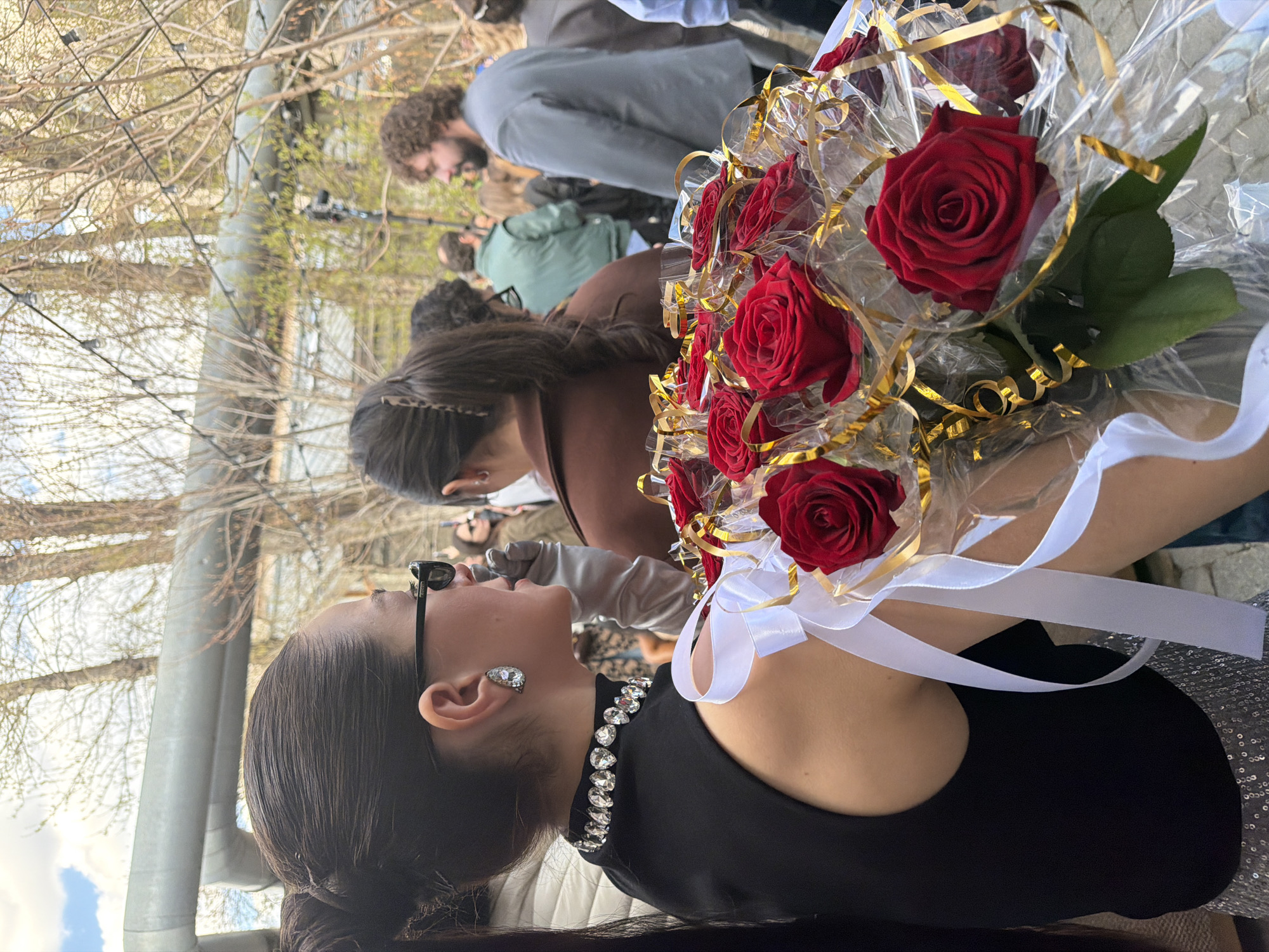 The image shows a woman in a black dress with a diamond necklace and earrings holding a bouquet of red roses wrapped in clear plastic with gold and white ribbons. She is wearing sunglasses and has her hair pulled back.
Behind the woman, there are several people standing. A man with a beard is visible in the background, wearing a grey shirt. There are also trees in the background.
Based on the people's attire and the bouquet, the scene might be at a celebratory event, such as a wedding. Due to lack of information, I cannot verify if the location is the city of Moscow, Russia.