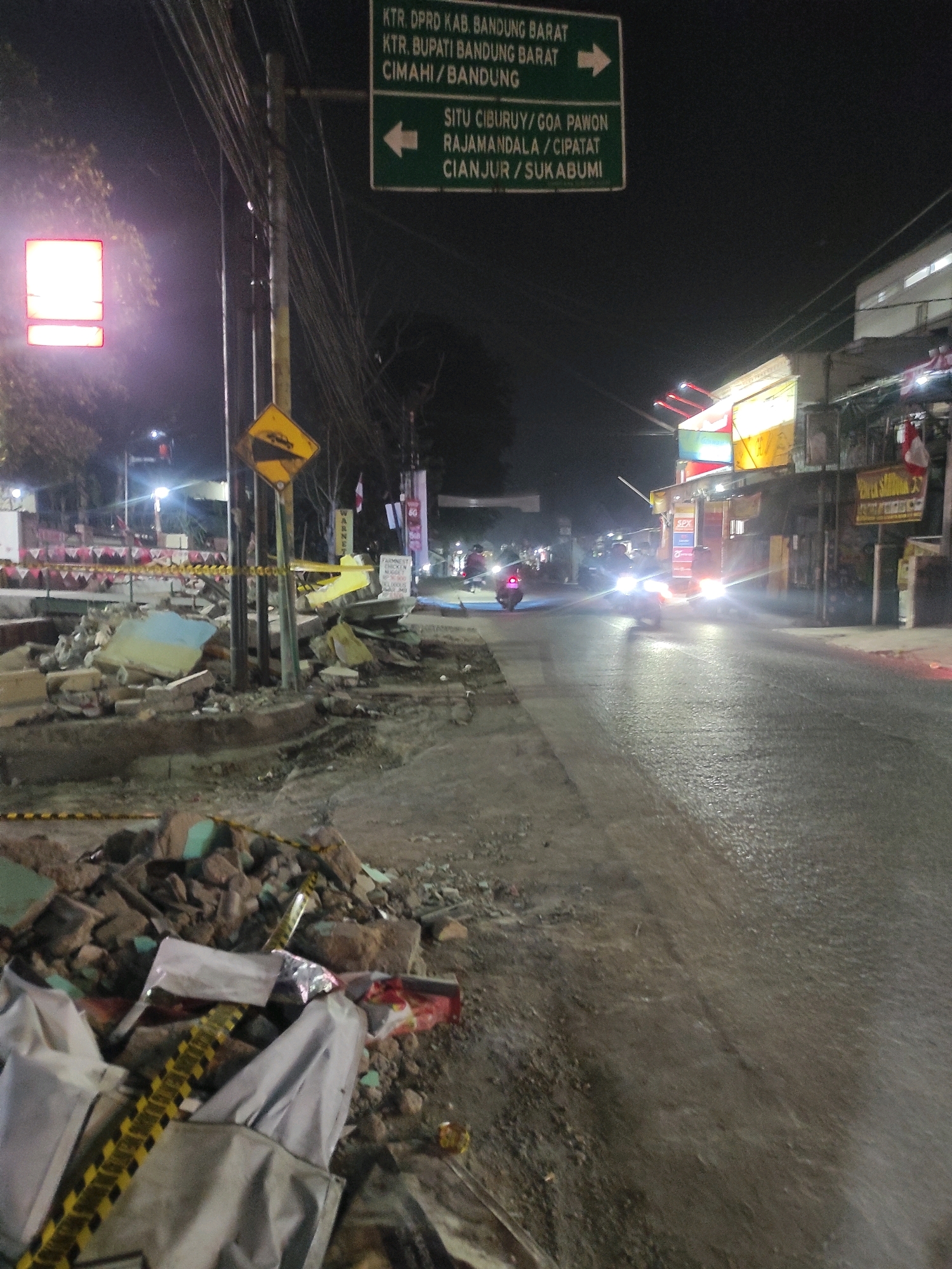 This is a night-time, medium-angle, full-frame, eye-level, outdoor, urban shot taken in Padalarang, Indonesia. The scene depicts a street with piles of rubble and debris lining the left side, suggesting recent demolition or construction. Yellow and black caution tape is visible, further indicating a construction or hazard zone.
In the foreground, a large pile of broken concrete, bricks, and what appears to be plastic sheeting or tarpaulin covers the ground. A yellow and black striped barrier tape is stretched across this debris. Further back along the left side of the street, more rubble is visible, interspersed with utility poles and streetlights. A red and white signpost with an arrow pointing left is also present.
The street itself is wet and reflects the bright headlights of approaching motorcycles. Two motorcycles are visible on the right side of the street, their headlights illuminating the wet asphalt. One motorcycle is in the mid-ground, and another is further in the distance. The street is lined with buildings and shops on the right side, some of which have illuminated signs. The sky above is dark.
A green directional signpost is prominently displayed in the upper left quadrant of the image, with text indicating destinations such as "KTR. DPRK KAB. BANDUNG BARAT", "KTR. BUPATI BANDUNG BARAT", and "CIMAHI / BANDUNG". Another sign below it indicates "SITU CIBURUY / GOA PAWON RAJAMANDALA / CIPATAT CIANJUR / SUKABUMI". A diamond-shaped yellow sign with a symbol of a steep incline is mounted on a pole near the rubble.