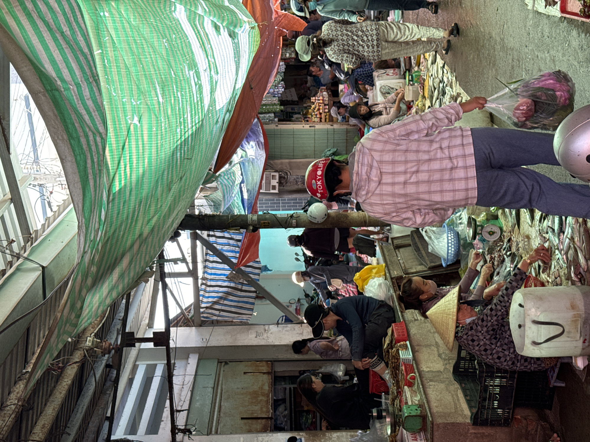 This image captures a bustling outdoor market scene in Mũi Né, Vietnam. The market is covered by a makeshift roof of green and white striped tarps, with some orange and red tarps also visible, creating a shaded environment. The ground appears to be paved.
In the foreground, a person wearing a pink plaid hoodie and blue pants is walking away from the camera, carrying a clear plastic bag filled with produce. A silver helmet is visible on the ground in front of them. To the left, a stall is set up with various goods laid out, including what appears to be dried fish. Several individuals are present at the stalls, some sitting and others working.
One woman, wearing a traditional conical hat, is sitting behind a stall and appears to be arranging fish. Another woman, wearing a mask and a patterned top, is also behind a stall, interacting with the produce. In the background, more people can be seen browsing and working, with motorcycles parked nearby, suggesting a typical day in a Vietnamese market. The overall atmosphere is one of lively commerce and daily life.