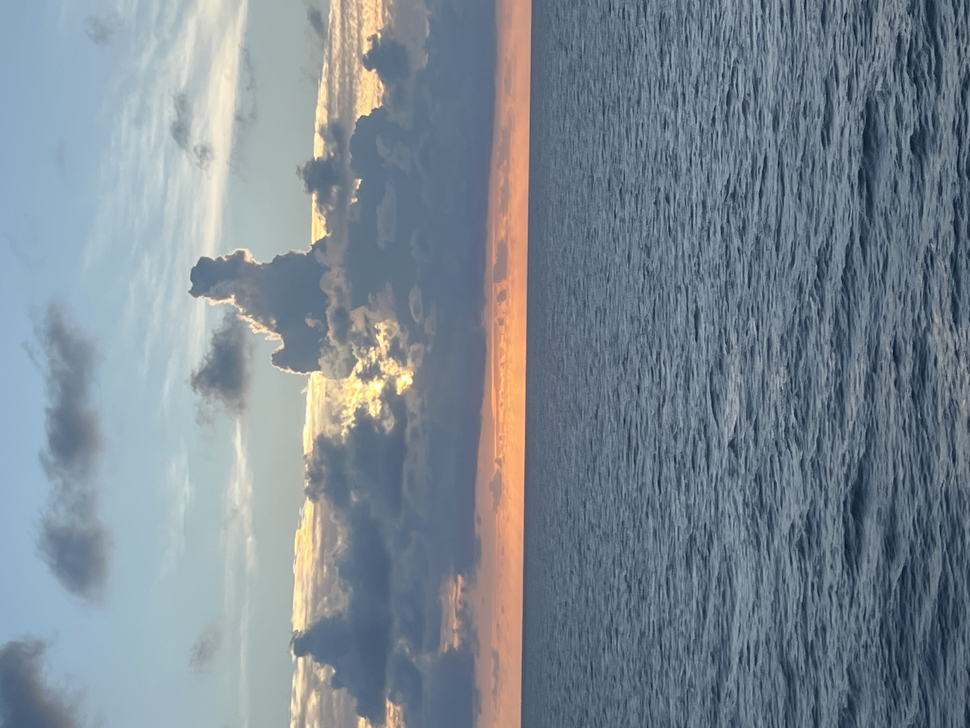 A wide shot captures a vast expanse of deep blue ocean meeting a horizon lined with the soft glow of sunset. The sky above is a canvas of pastel hues, transitioning from a warm orange near the horizon to cooler blues and grays as it ascends. Dark, voluminous clouds dominate the upper portion of the scene, their edges illuminated by the setting sun, creating a dramatic contrast. A particularly striking cloud formation stands out, shaped uncannily like a standing figure. The ocean surface is rippled, reflecting the colors of the sky and hinting at a gentle breeze. The overall impression is one of serene natural beauty, with the cloud formation adding a touch of whimsical wonder to the otherwise tranquil seascape. The image is taken at an eye-level perspective, with no people or identifiable man-made structures present.