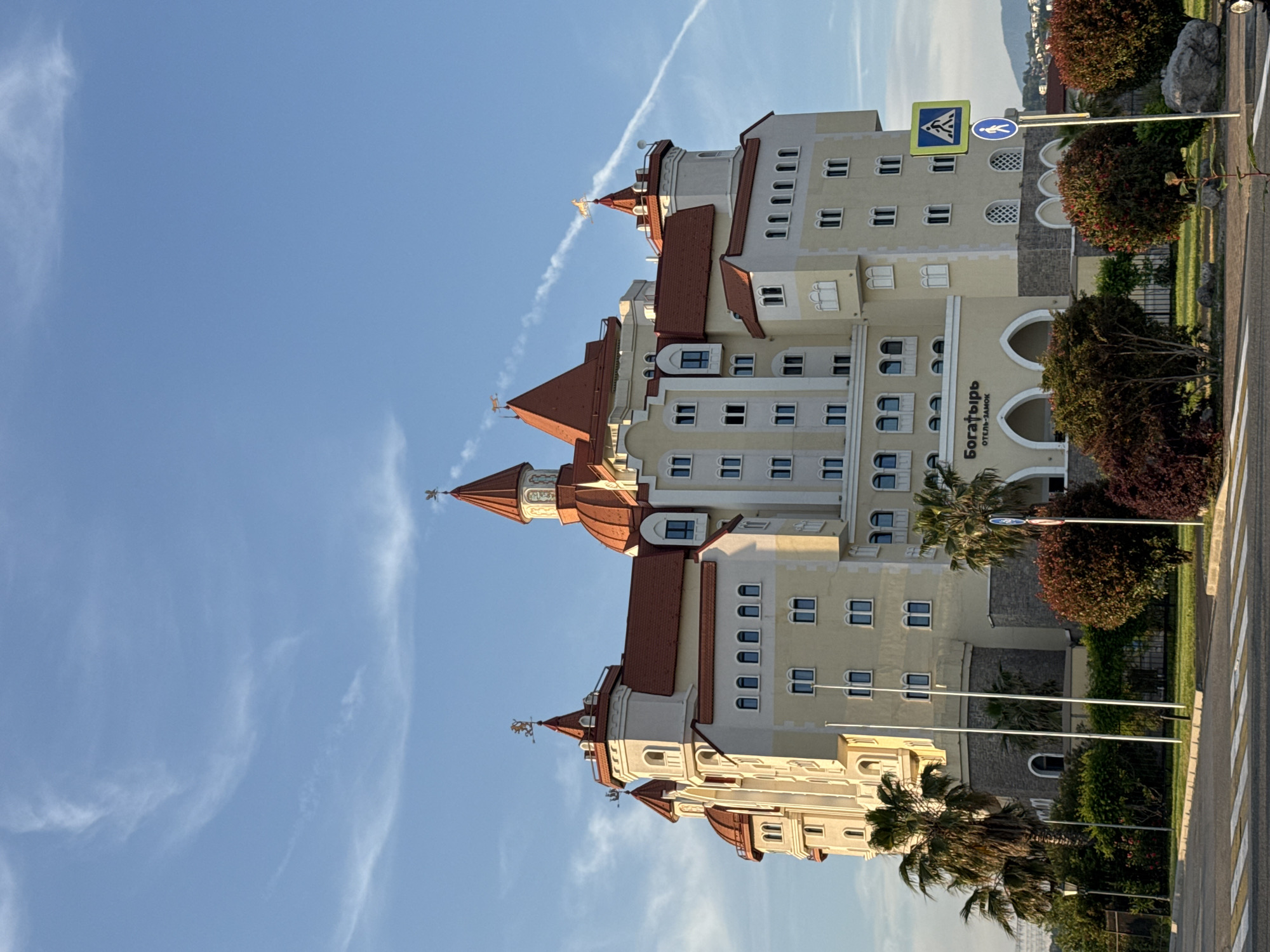 The image presents the Bogatyr Hotel-Castle located in Sirius, Russia, under a clear blue sky with a prominent contrail extending from the upper right. The multi-story building, designed in a fairytale castle style, features a light cream-colored facade with numerous arched and rectangular windows. It is crowned with distinctive reddish-orange pitched and domed roofs, with several spires topped by golden weathervanes depicting mythical creatures, including one resembling a griffin and another a horse. The base of the building is constructed with a darker, stone-like material. An arched entrance on the ground floor displays the hotel's name in Cyrillic, "Богатырь ОТЕЛЬ-ЗАМОК".
The foreground includes a pedestrian crosswalk marked with white stripes. Various green and reddish-brown bushes are strategically planted, alongside several tall palm trees on the left. Three white, vertical poles, possibly flagpoles, stand in the mid-left. On the right, two blue and white pedestrian street signs are mounted on a single pole; the upper sign warns of a pedestrian crossing, and the lower indicates a pedestrian path. A dark street lamp is visible on the far left. The scene is illuminated by warm, directional sunlight, suggesting either early morning or late afternoon. No individuals are present, and no actions or interactions are depicted.