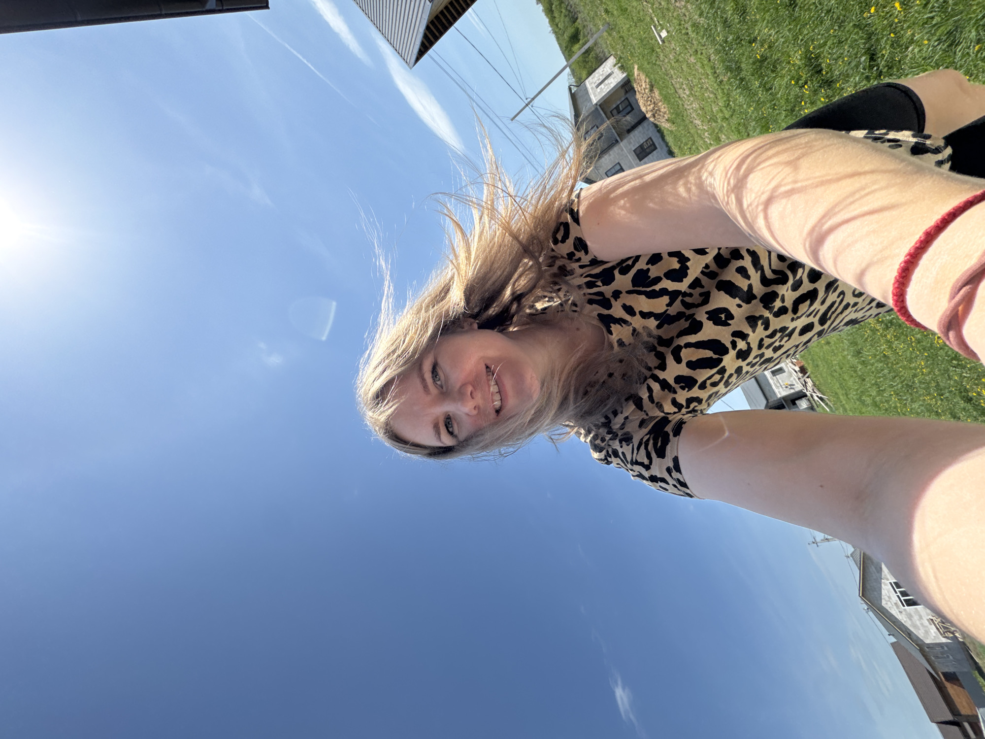 A low-angle selfie of a young woman with blonde hair and a leopard print dress. She is smiling. Her left arm is visible, with a red bracelet on her wrist. The background shows a bright blue sky with some contrails, buildings, and grass with yellow flowers. The location is in Ферма, Russia.