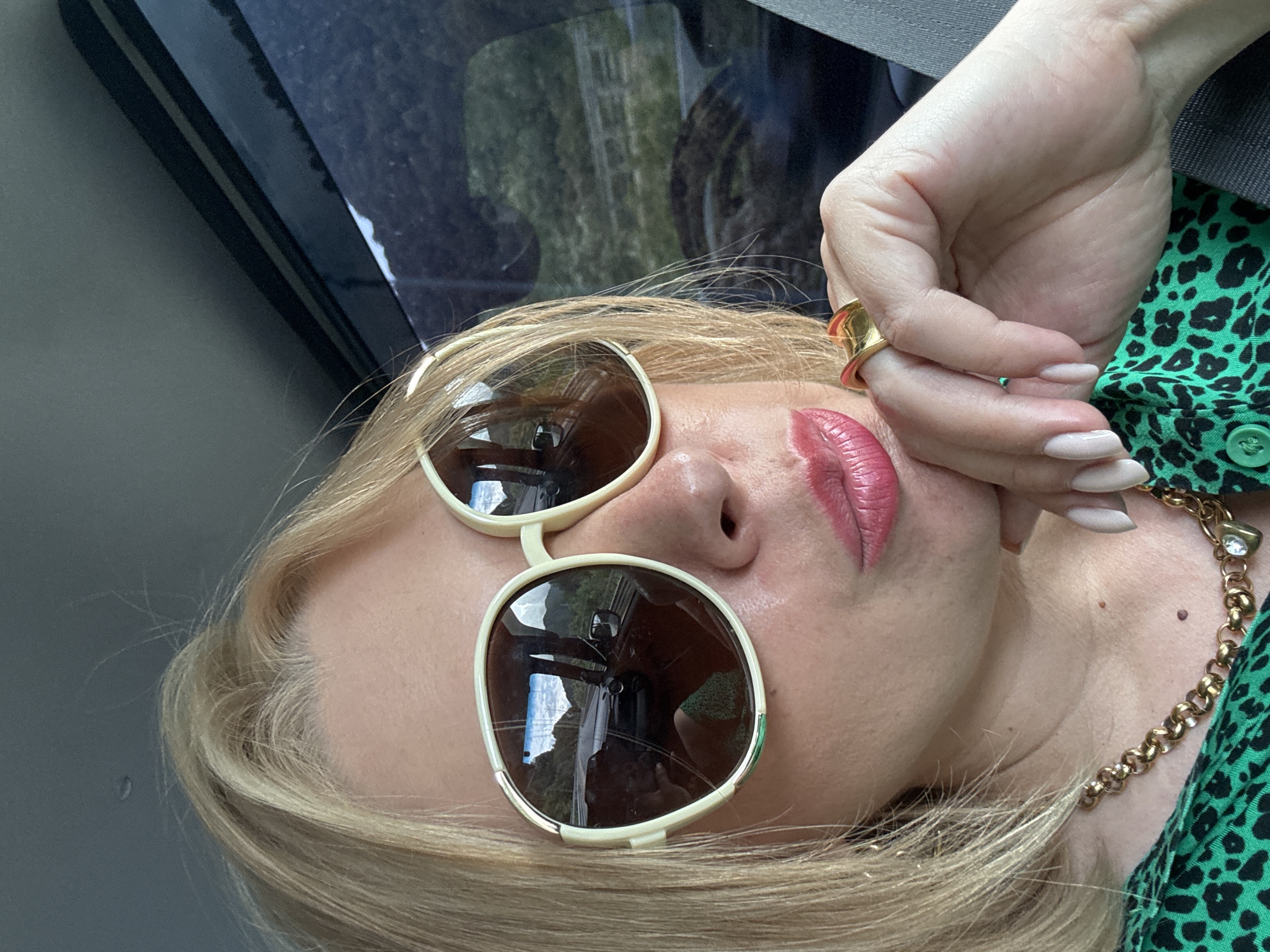 A close-up, selfie-style shot captures a fair-skinned woman with blonde hair and light skin tone, wearing large, cream-rimmed sunglasses with brown lenses. Her lips are a bright pink, and she is wearing a gold necklace with a heart-shaped pendant and a large, abstract gold ring on her right hand, which is resting on her chin. She is also wearing a green top with a black leopard print. The background appears to be the interior of a vehicle, with reflections of a road and mountains visible in the sunglasses. The scene is set in the city of Kepsha, Russia.