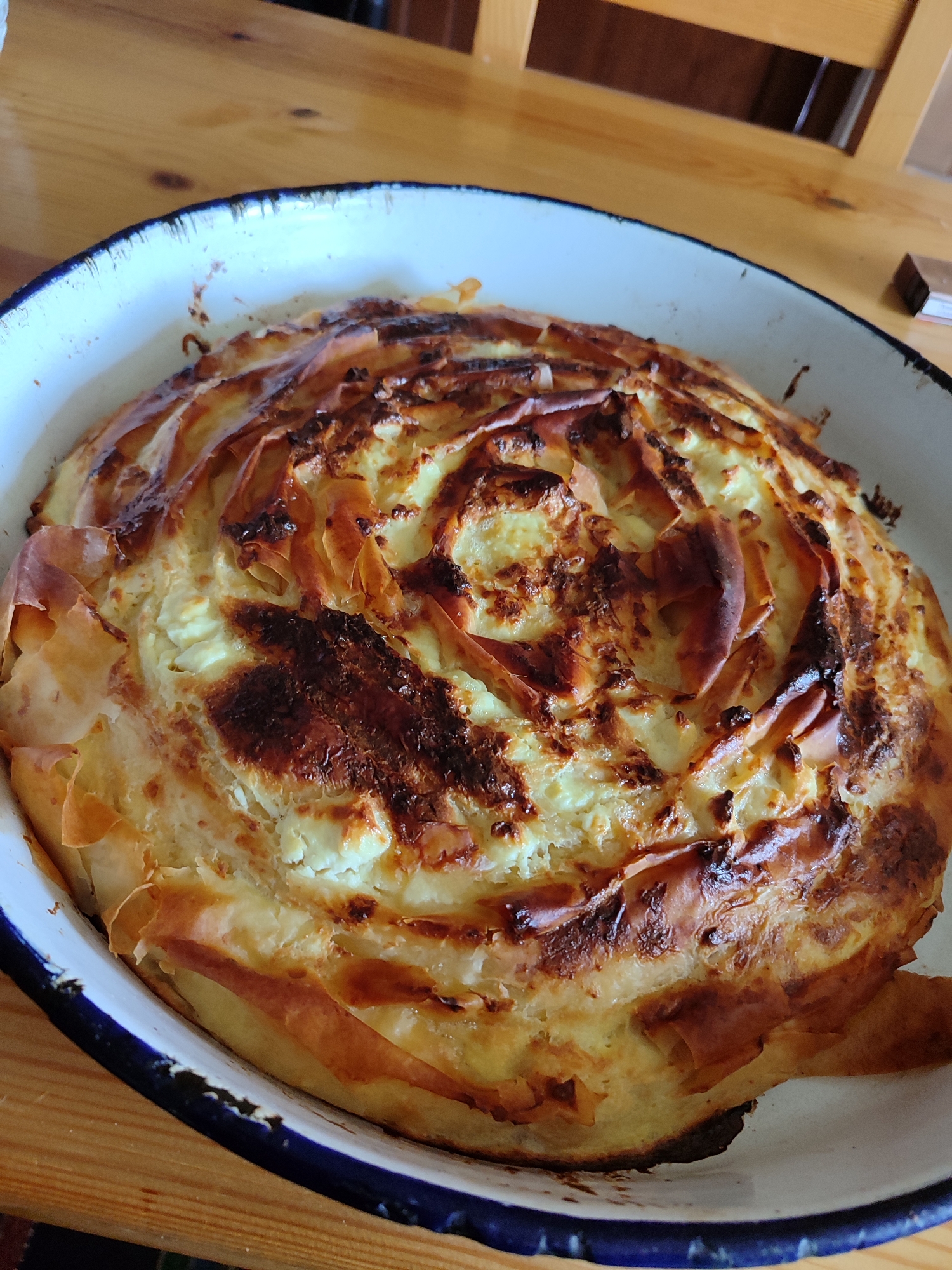 A close-up shot shows a baked cheese phyllo pie, likely homemade, nestled in a white enamel baking dish with a blue rim. The pie exhibits a circular design with flaky, golden-brown phyllo pastry arranged in layers. The filling appears creamy and light in color, possibly a cheese mixture. The top of the pie is darker in some areas, indicating varied browning or slightly burnt edges. The baking dish is situated on a wooden table. This image was taken from the city of Varshilo, Bulgaria.