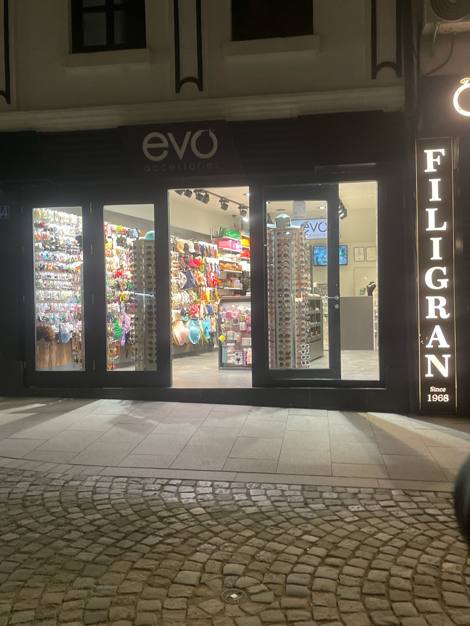The image shows the storefront of "evo accessories" in Prizren, Kosovo, at night. The store has a dark facade with illuminated windows showcasing a variety of accessories, including sunglasses, bags, and jewelry. To the right of the store entrance, a vertical sign reads "FILIGRAN SINCE 1968" in illuminated white lettering. The street in front of the store is paved with cobblestones and large tiles. The overall impression is that of a well-lit retail establishment in an urban setting.