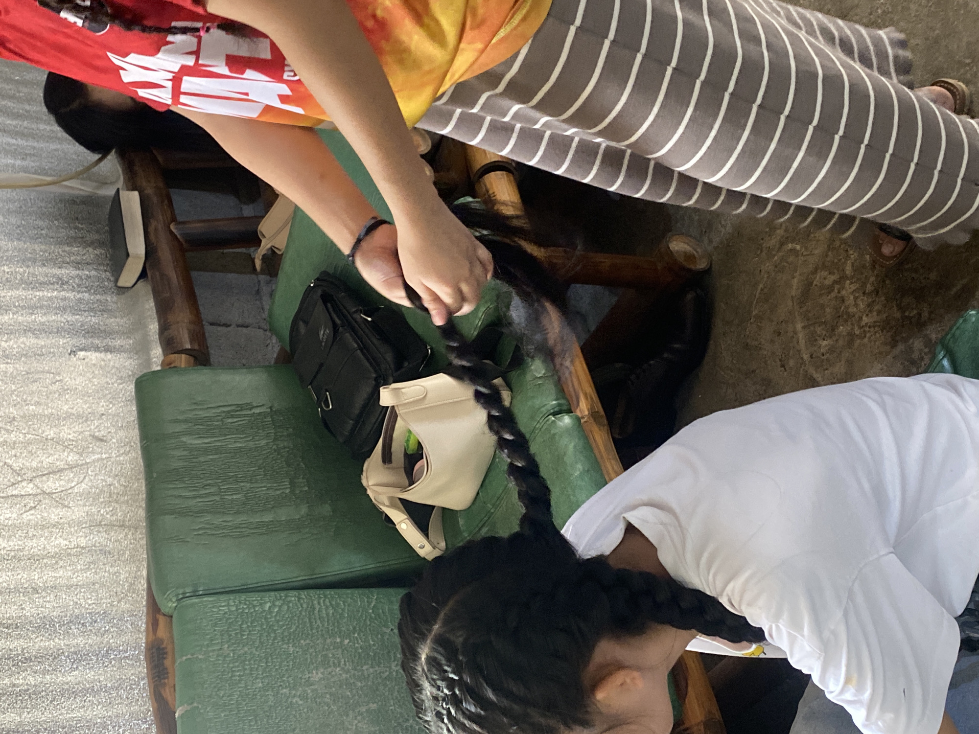 In an indoor setting in Davao City, Philippines, a person with long black hair is having their hair braided by another person. The person receiving the braid is wearing a white t-shirt and their hair is already styled in two thick braids that extend down their back. The person doing the braiding, wearing a red and orange tie-dye shirt and a grey and white striped skirt, is holding a section of hair and actively braiding it.
The scene appears to be in a casual, perhaps outdoor or semi-outdoor, area. To the left of the frame, there are green upholstered benches, likely part of a seating area. On the bench behind the person receiving the braid, a black bag and a small cream-colored purse are visible. In the background, a textured metallic wall and wooden furniture can be seen. A small book is also placed on a wooden surface in the background. The flooring is a rough concrete surface. The overall impression is one of a relaxed, everyday activity taking place in a simple environment.